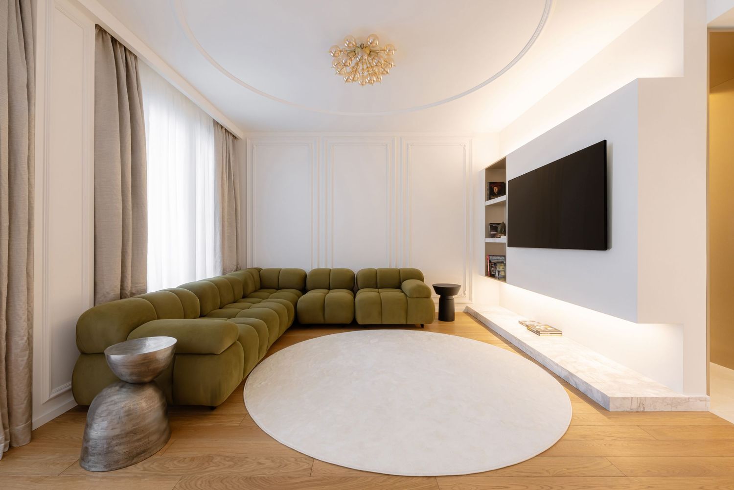 Modern living room with olive green sectional sofa, wall-mounted TV, round white rug and wooden flooring.