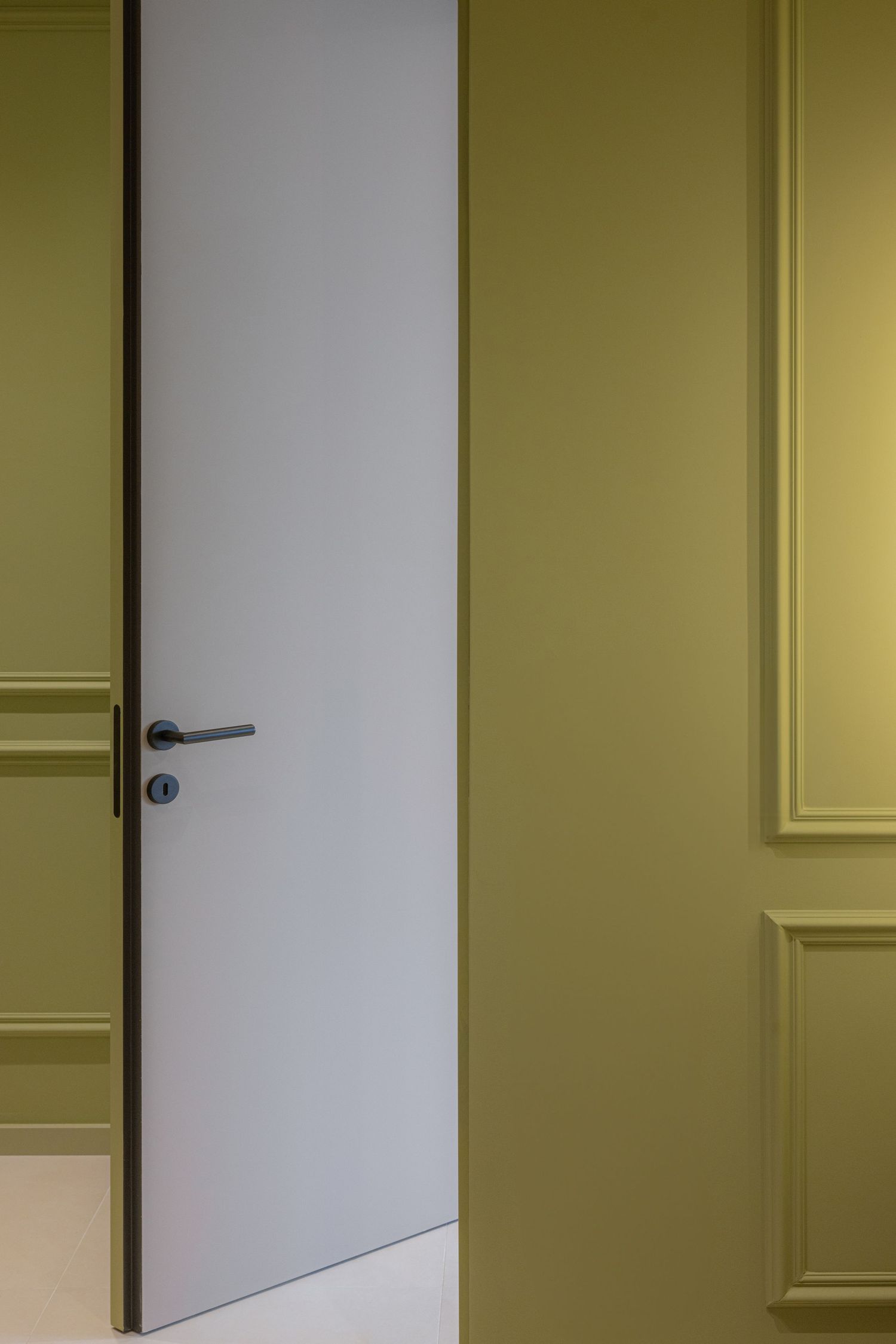 White interior door partially open in a hallway with yellow-tinted lighting and white trim molding by Vedrana Markovic.