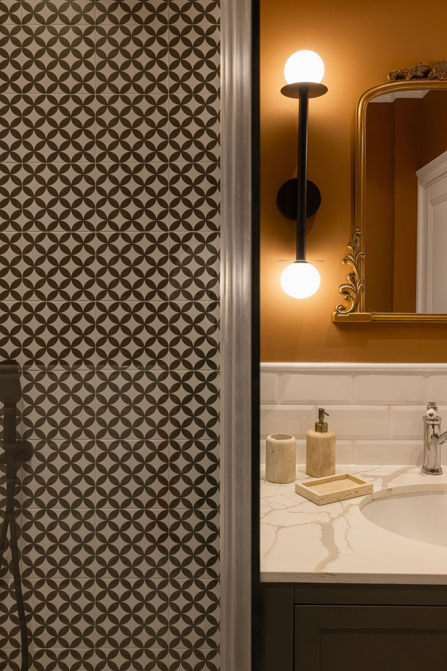 A modern bathroom interior with geometric patterned wallpaper and warm lighting fixtures mounted above a white vanity by Vedrana Markovic.