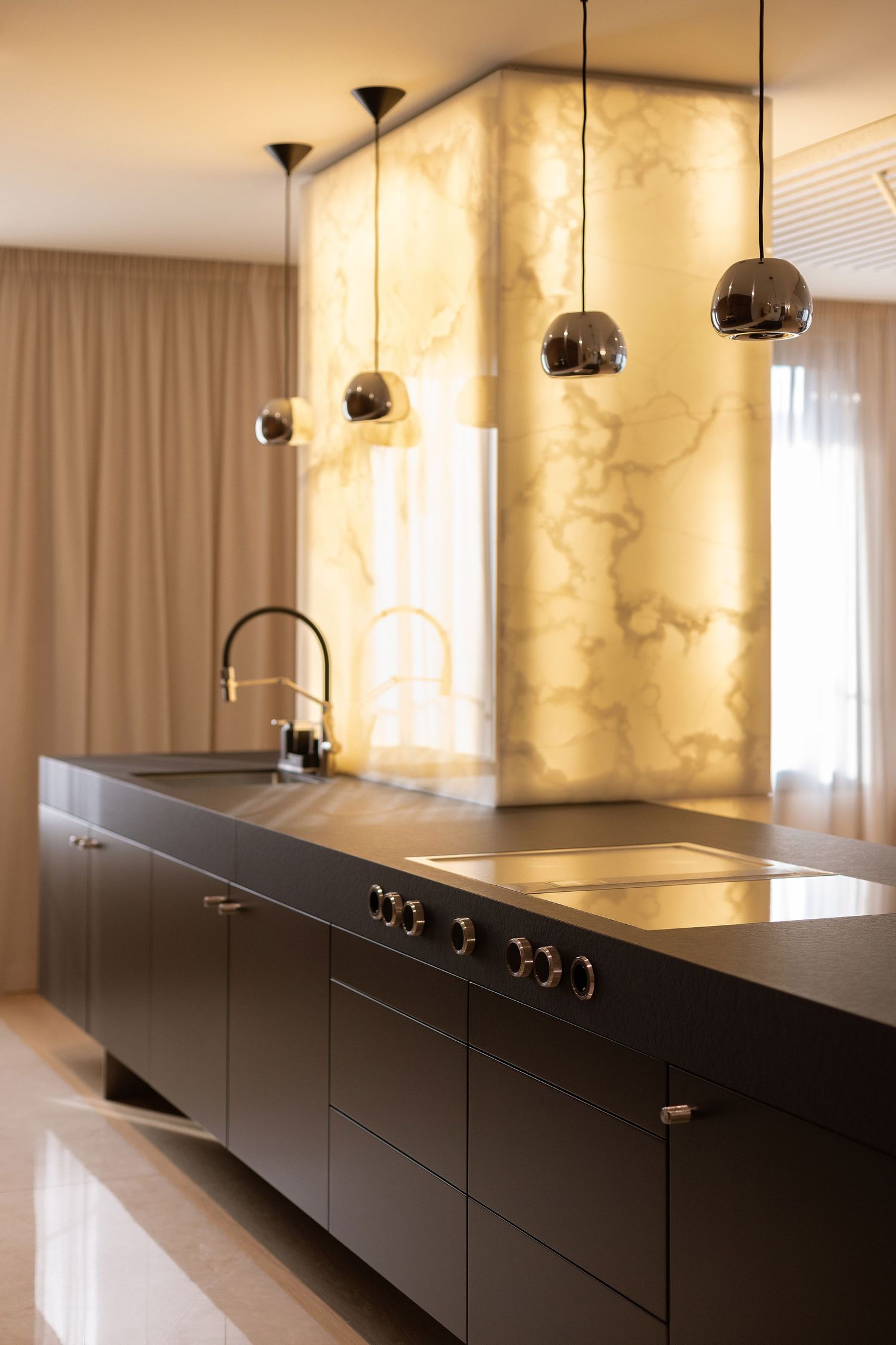 Modern kitchen interior with illuminated marble backsplash and sleek black cabinetry features pendant lighting fixtures by Studio Fluid.