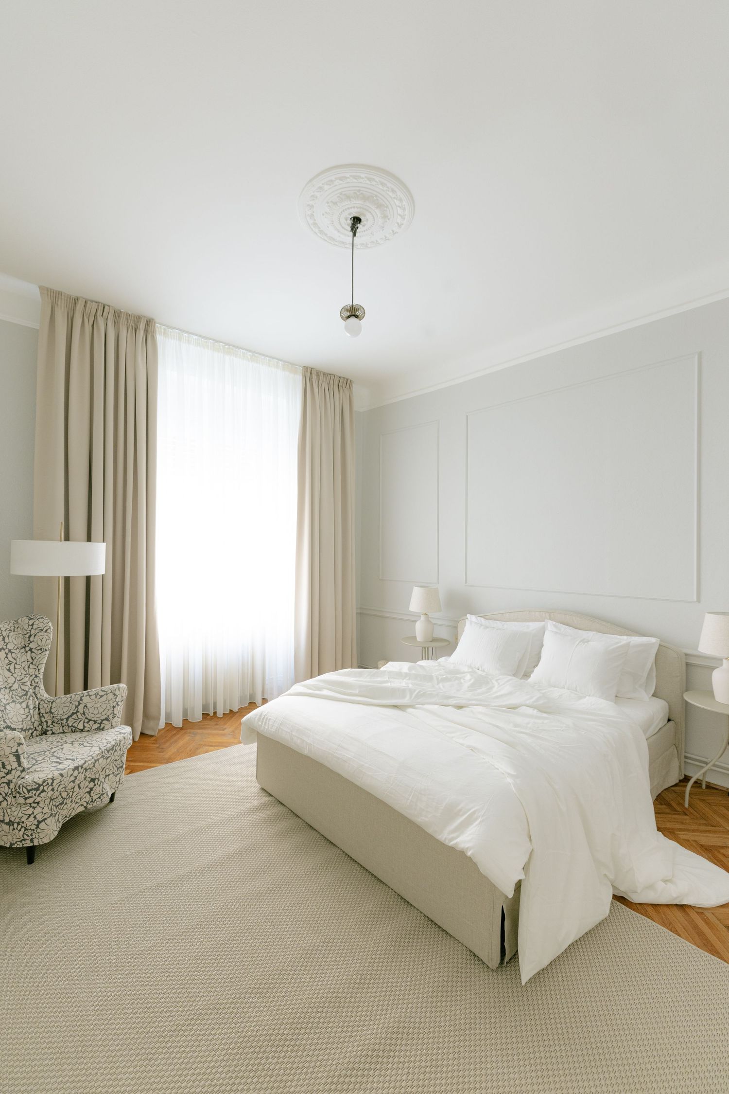 Elegant white bedroom with a large bed, beige curtains, patterned armchair, and ornate ceiling medallion.