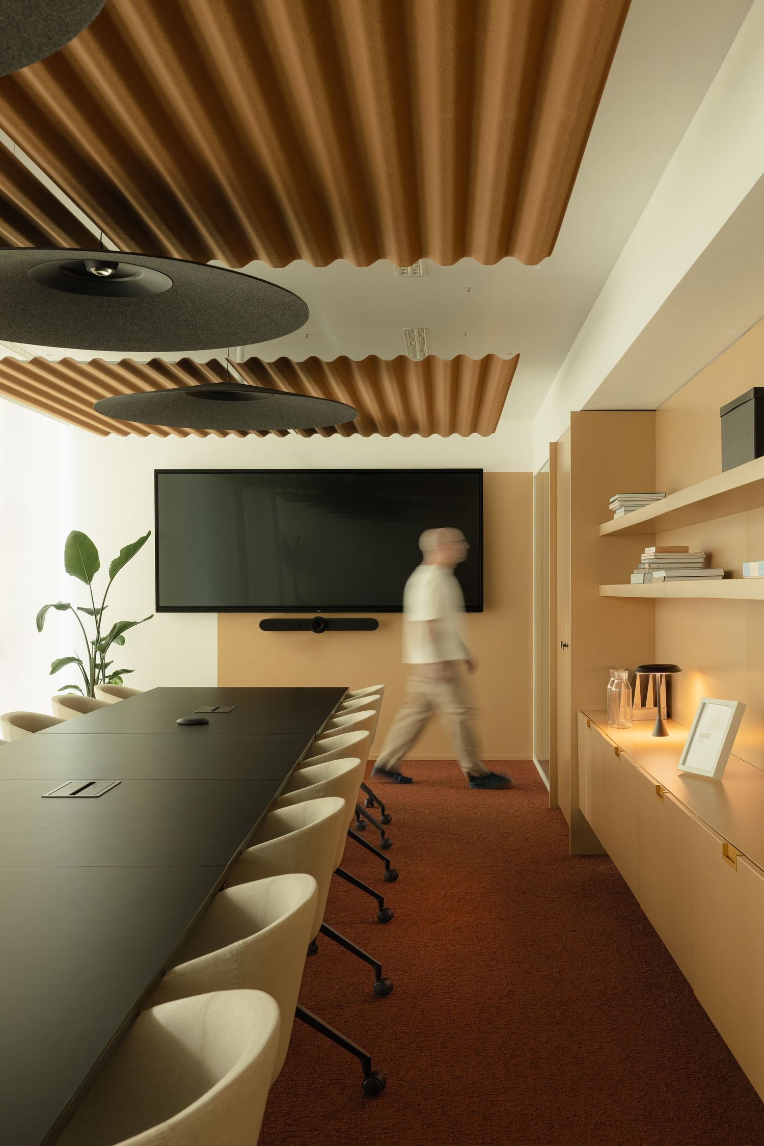 Modern conference room with long dark table, cream chairs, wood slat ceiling, wall-mounted TV, and built-in shelving.
