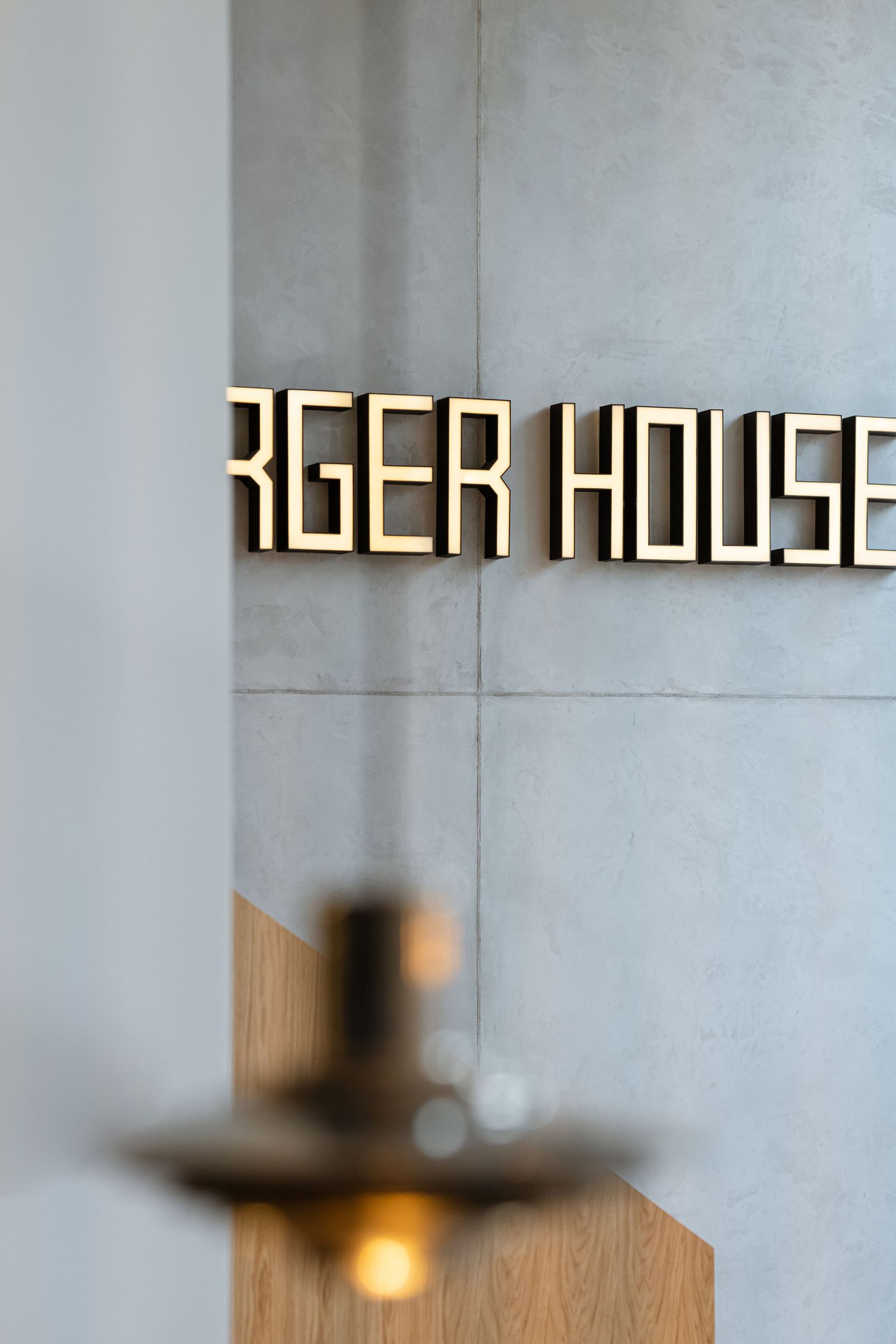 Modern illuminated 'BURGER HOUSE' signage mounted on a gray wall with wooden accent and blurred foreground detail by Studio Fluid.