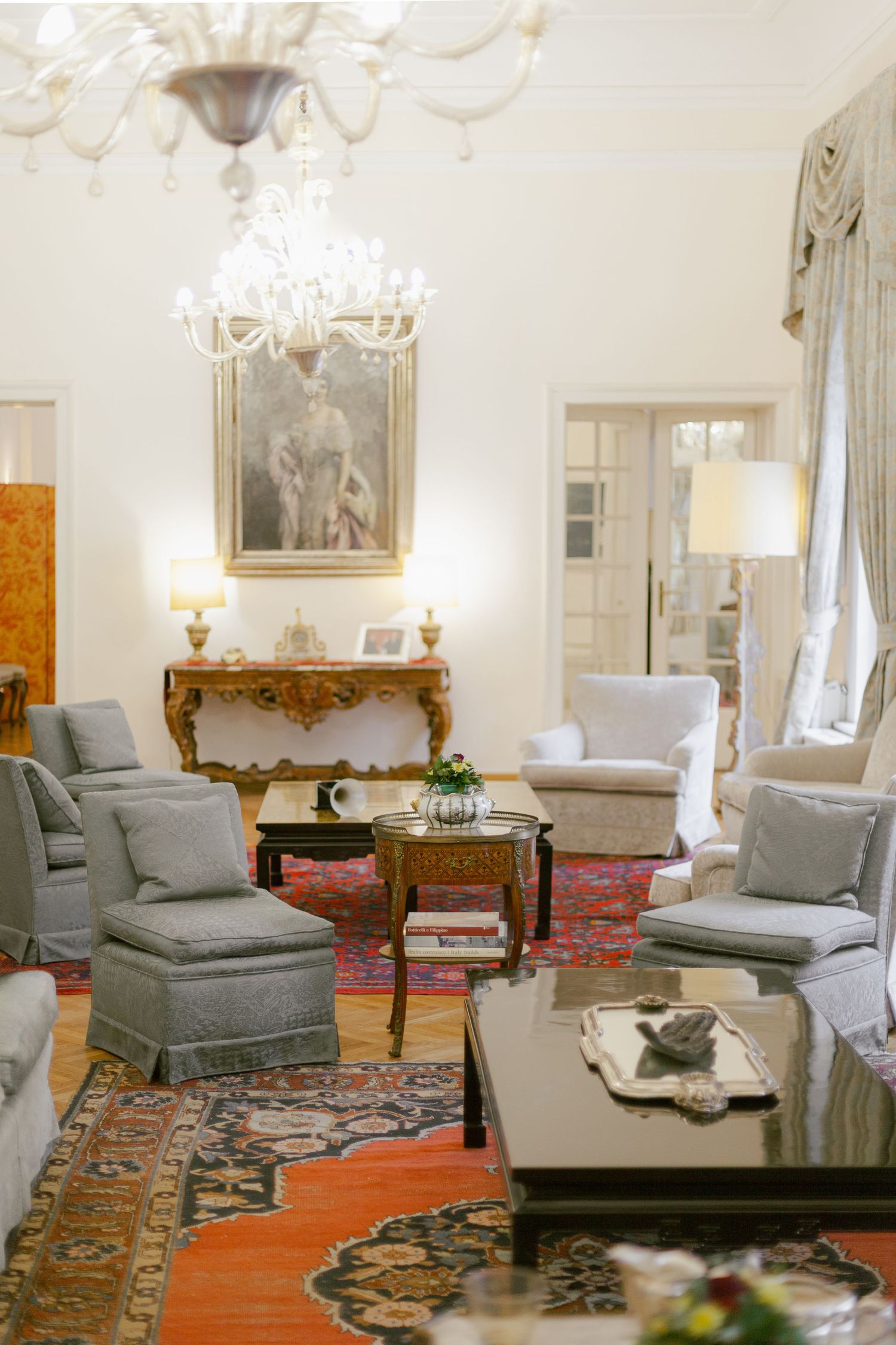 Elegant living room with crystal chandelier, ornate console table, gray upholstered chairs and Persian rug on hardwood floor.
