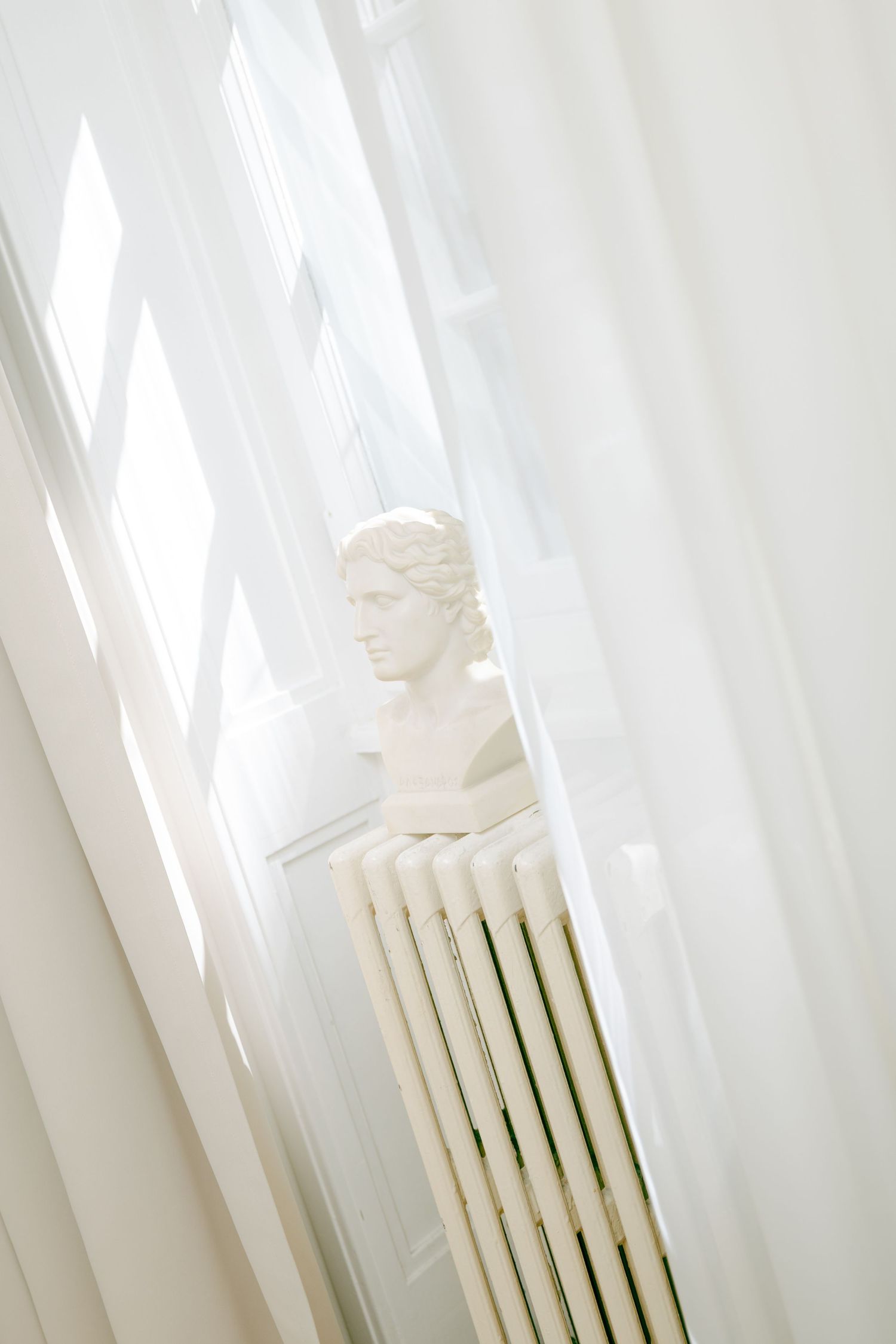 White classical bust sculpture on a radiator beside sheer curtains in a bright, airy room.