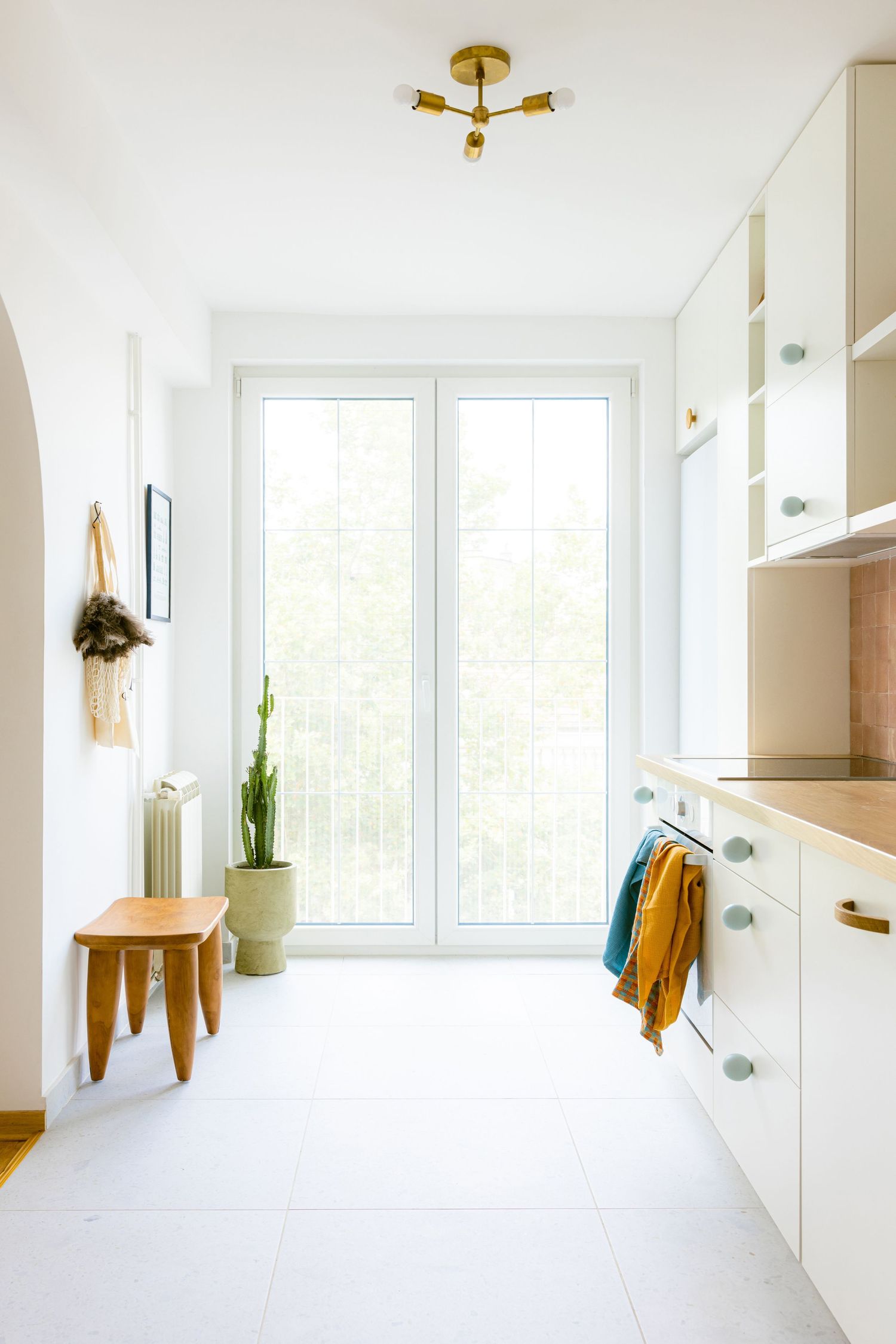 Bright white minimalist kitchen interior with gold light fixtures, white cabinets, and natural light from windows by Maja Interiors.