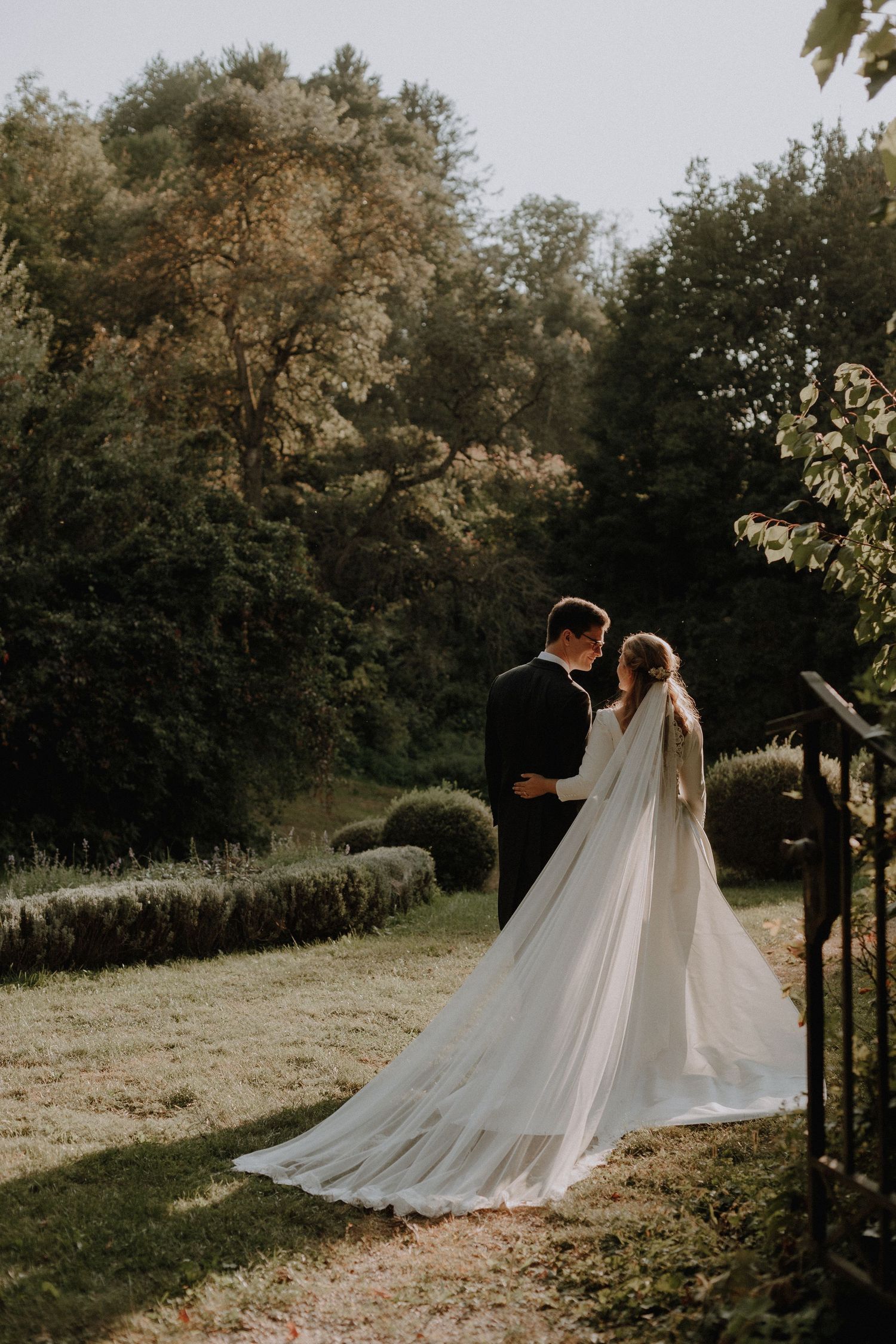 A romantic wedding couple in a scenic outdoor setting with a flowing white dress and long veil in golden sunlight.