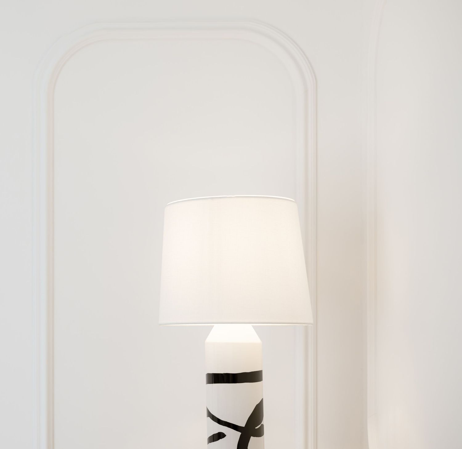 White table lamp with black accents casting soft light against a plain white wall in a minimalist setting.