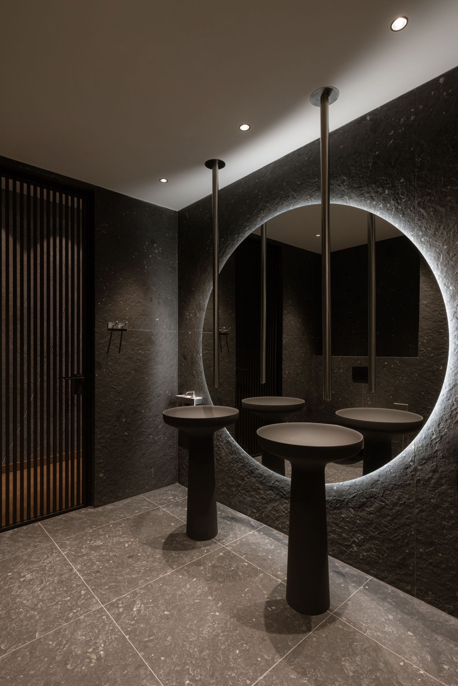 Modern dark bathroom interior with circular backlit mirror and pedestal sinks featuring dramatic pendant lighting by Aleksandar Savikin.