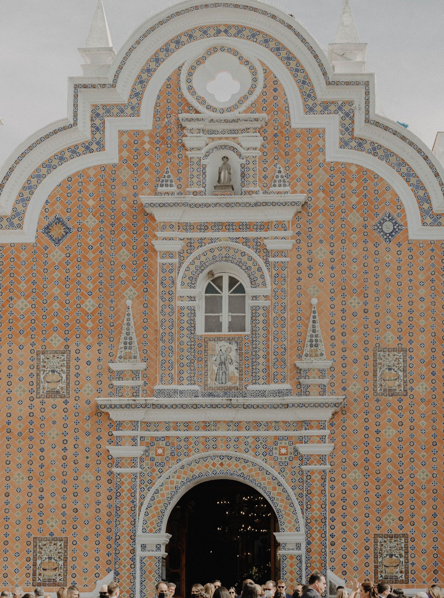 A large group gathers in front of an ornate brick and stone building with decorative arched architecture.