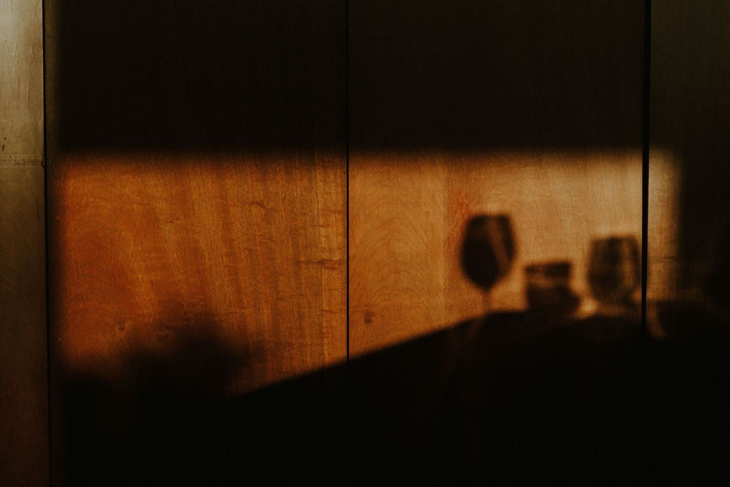 A dark and moody bar counter with wine glasses and bottles silhouetted against warm lighting.