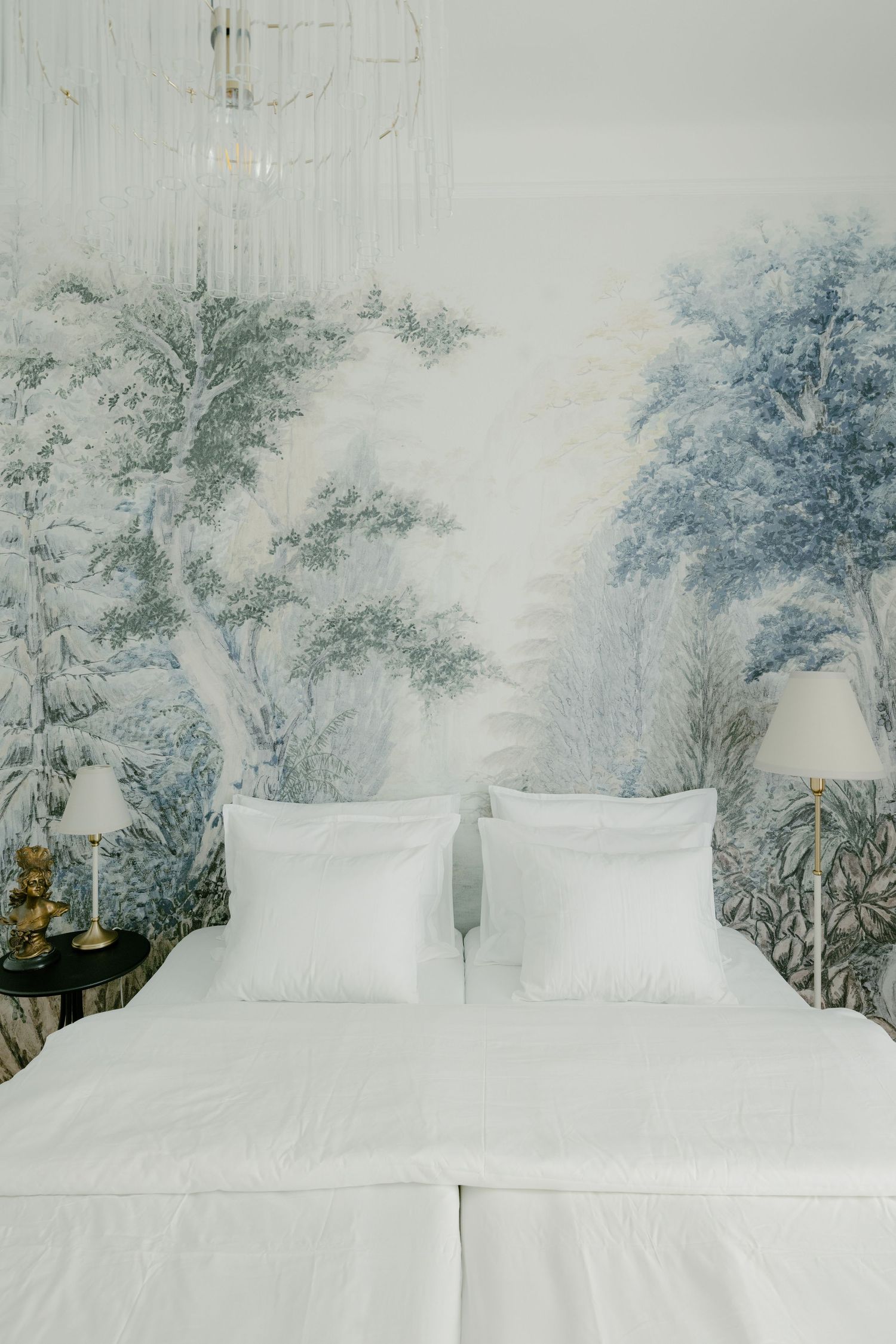 Elegant bedroom with white bedding and blue-green toile forest mural wallpaper, flanked by gold table lamps.
