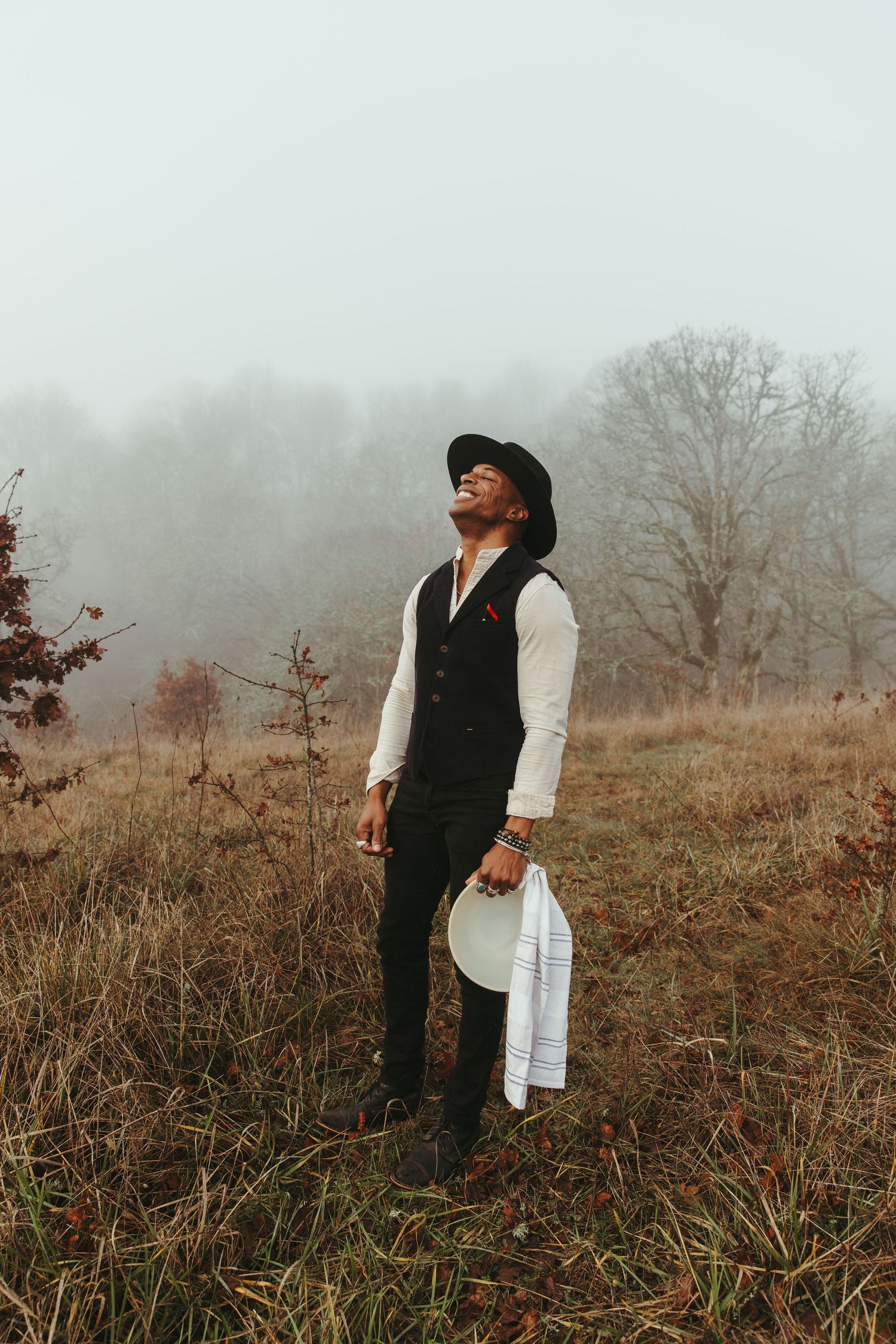 A stylish man in a black vest and hat stands in a misty field, holding a white cloth, surrounded by autumn foliage.