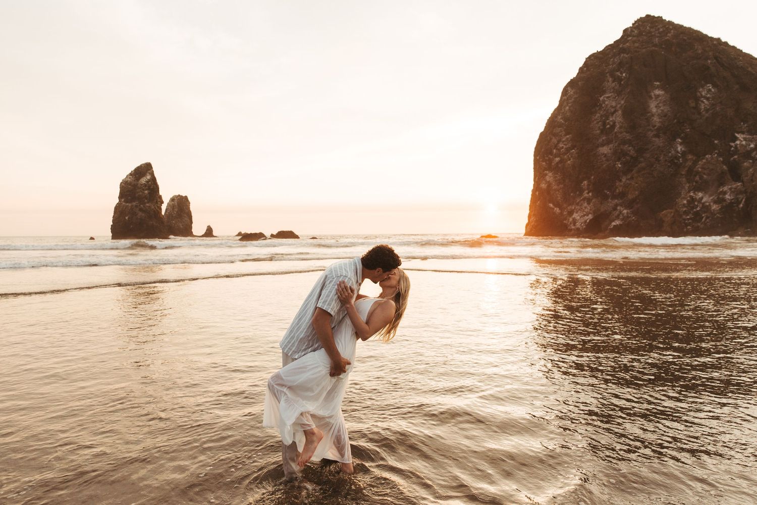Couple embraces on rocky beach during golden sunset with ocean waves and sea stacks in background.