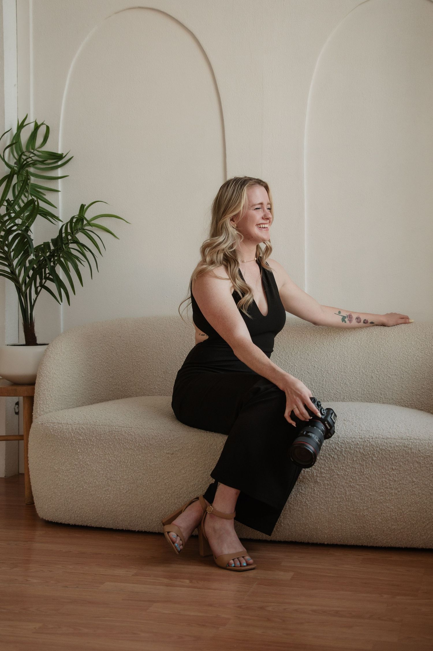 A person in a black dress sits casually on a beige couch in a minimalist room with hardwood floors and a potted plant.