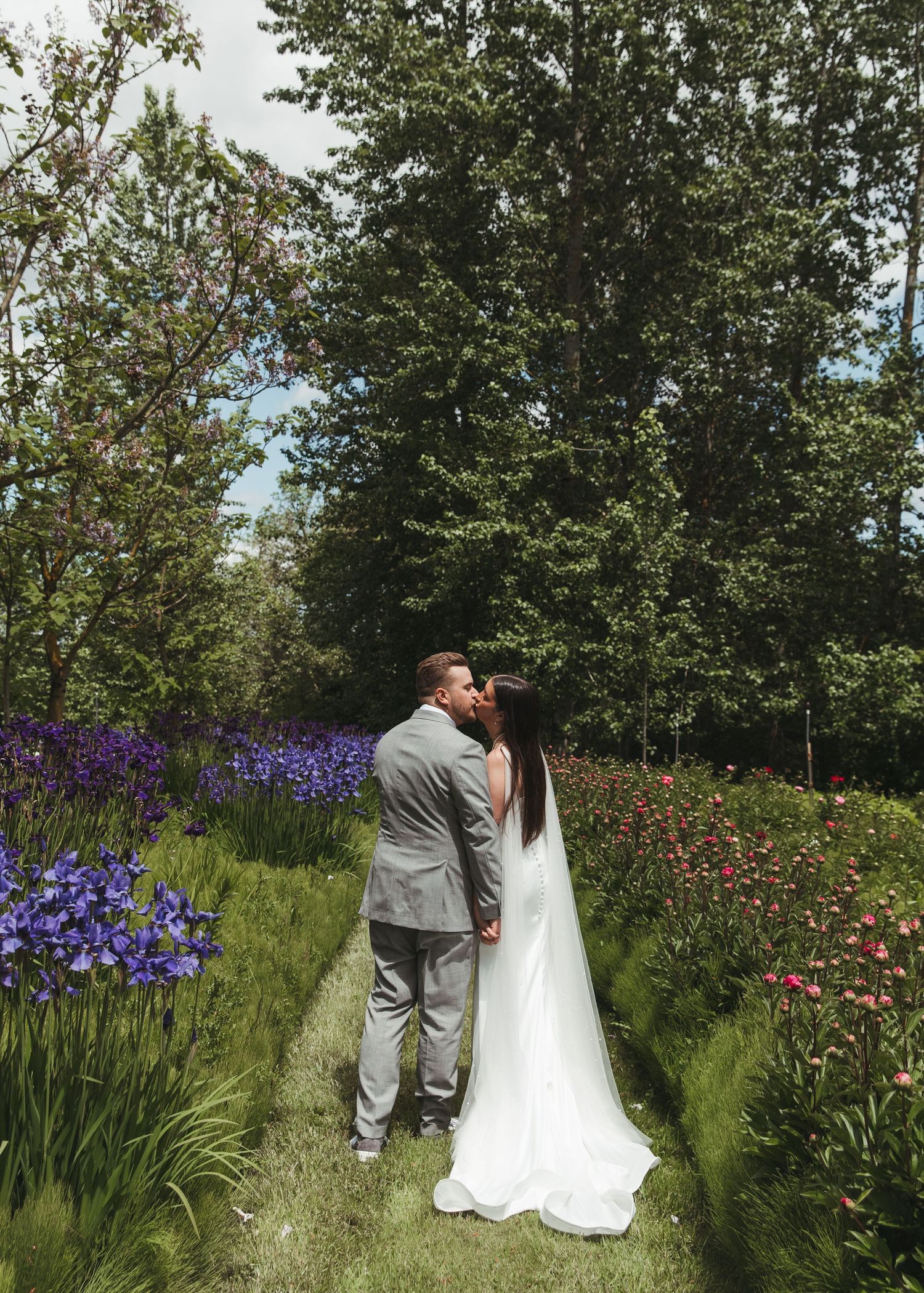 Bride and groom kissing in a lush garden path surrounded by purple and pink flowers on their wedding day.