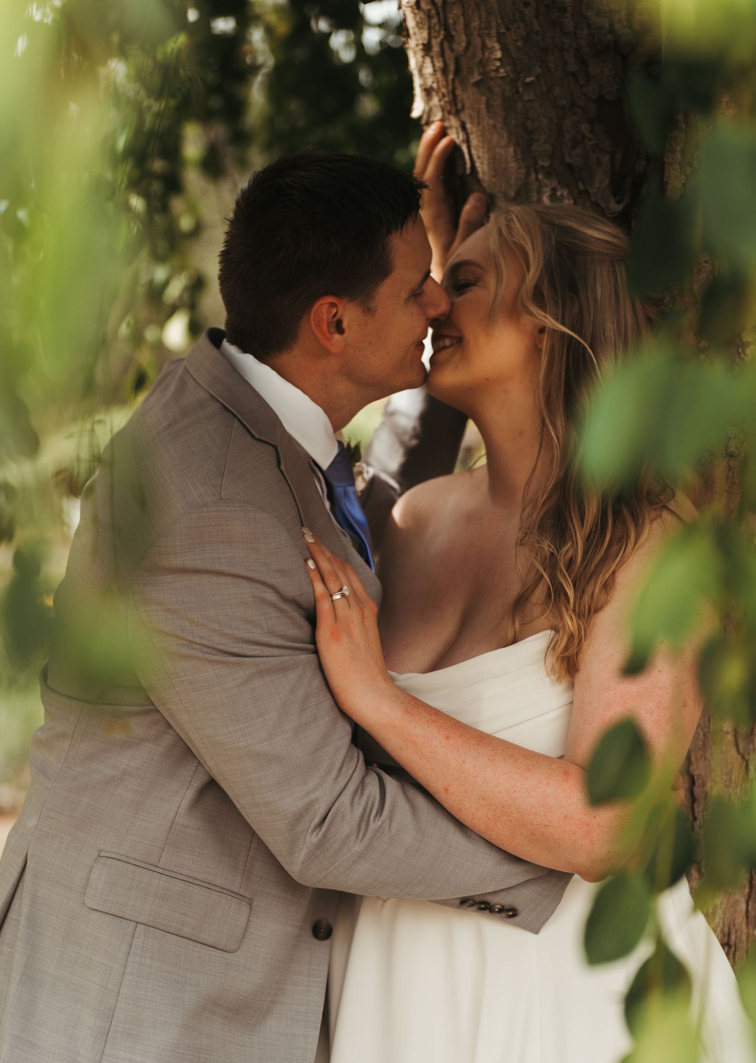 A bride and groom share a romantic almost-kiss beneath lush green trees in an outdoor wedding portrait.