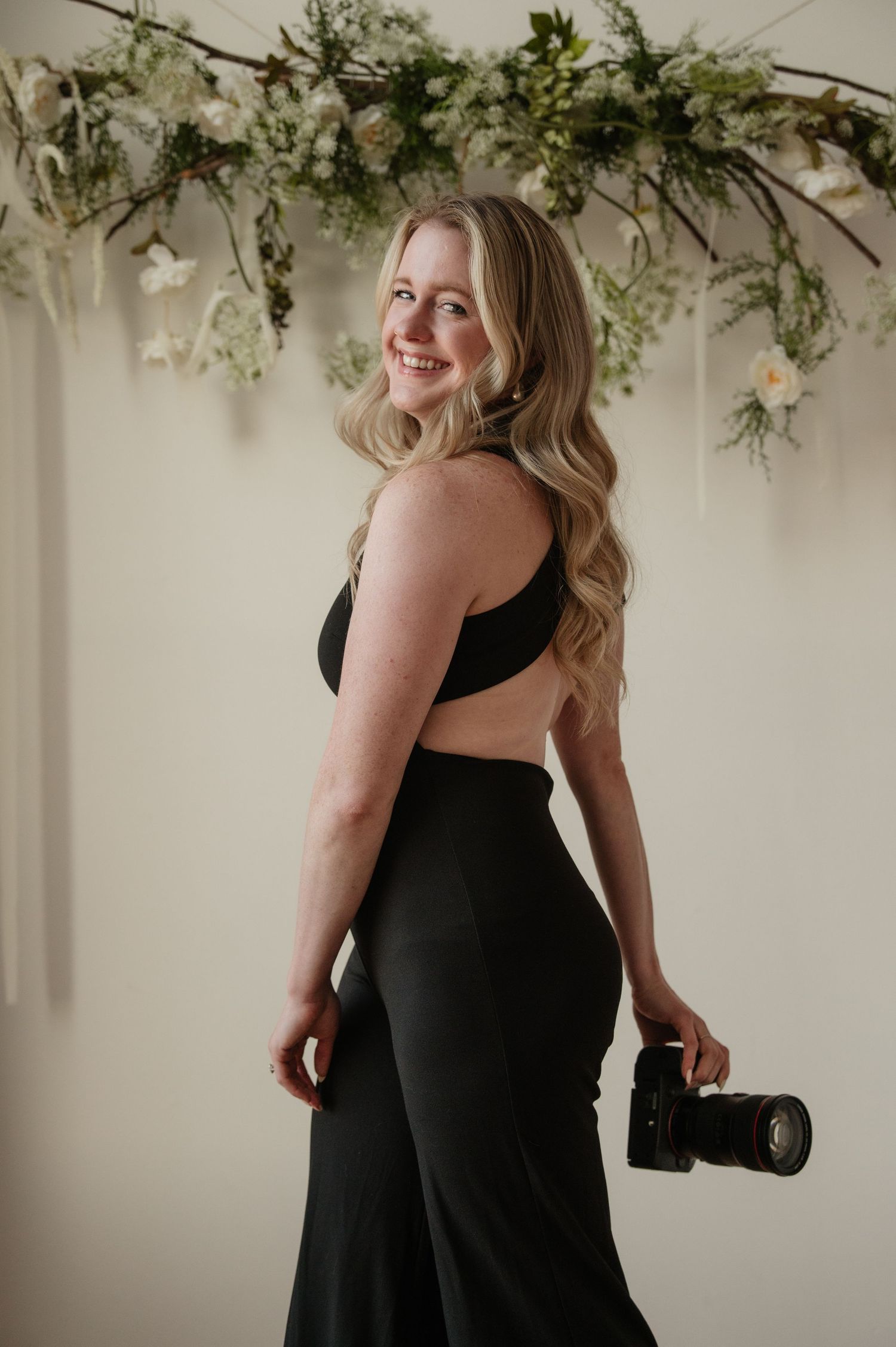 Professional studio portrait session featuring artistic greenery decorations and a classic black dress in light cream backdrop.