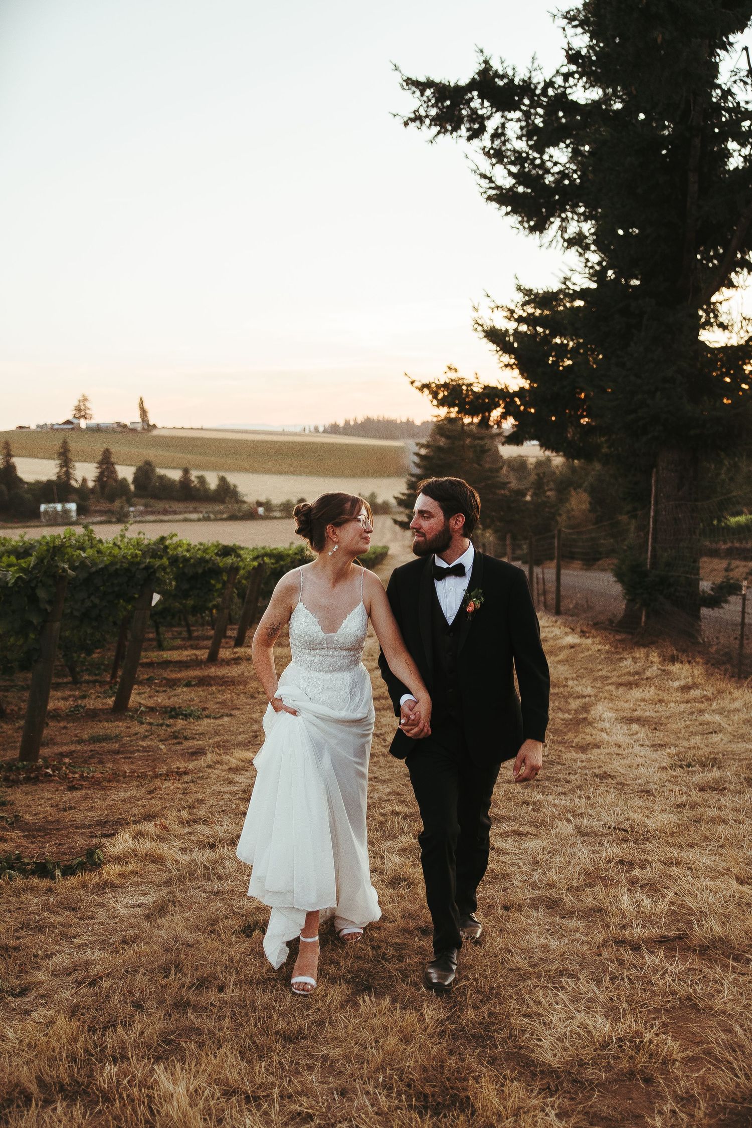Bride and groom walk hand in hand through vineyard at golden hour, she in white gown, he in black tuxedo.