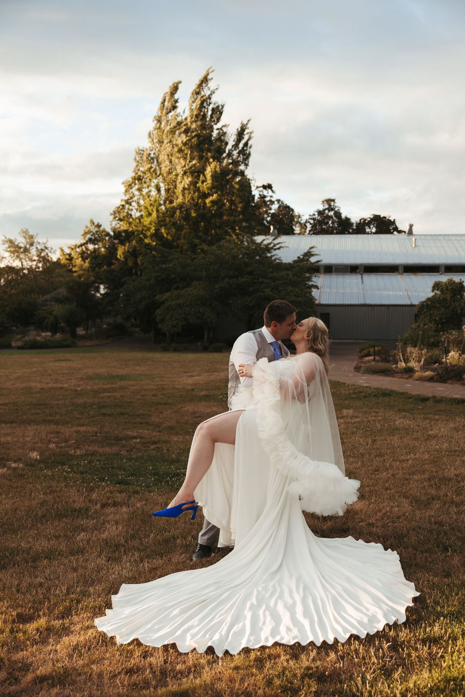 Groom lifts bride in white gown and blue heels, sharing a kiss outdoors at golden hour near a rustic barn.
