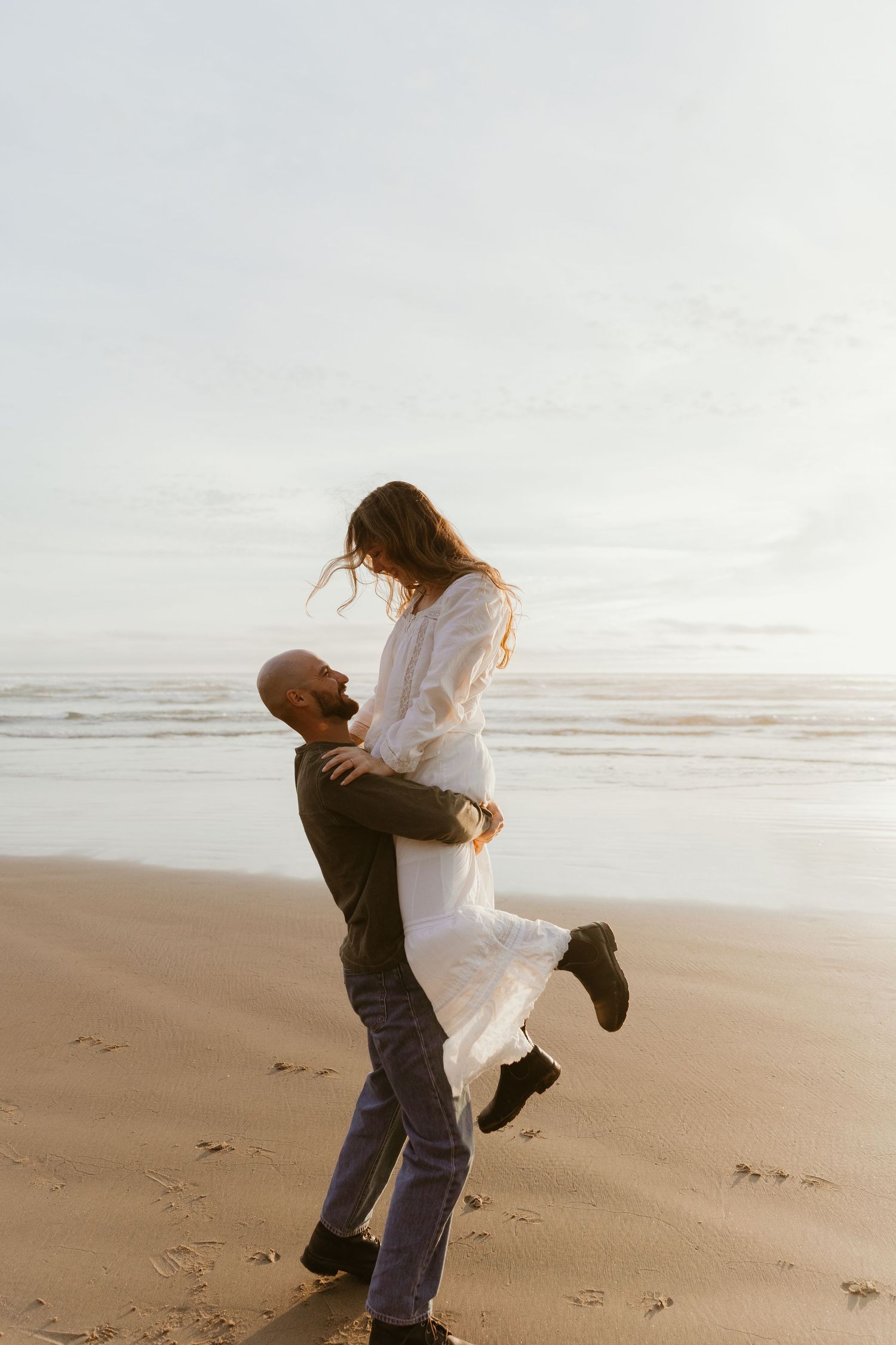A man lifts a woman in a white dress on a sandy beach at sunset, waves in the background.