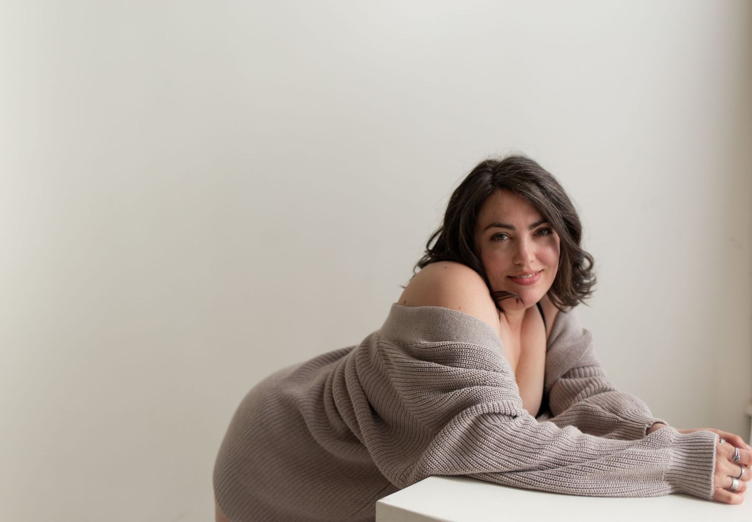 Cozy boudoir portrait of someone in an oversized sweater against white wall.