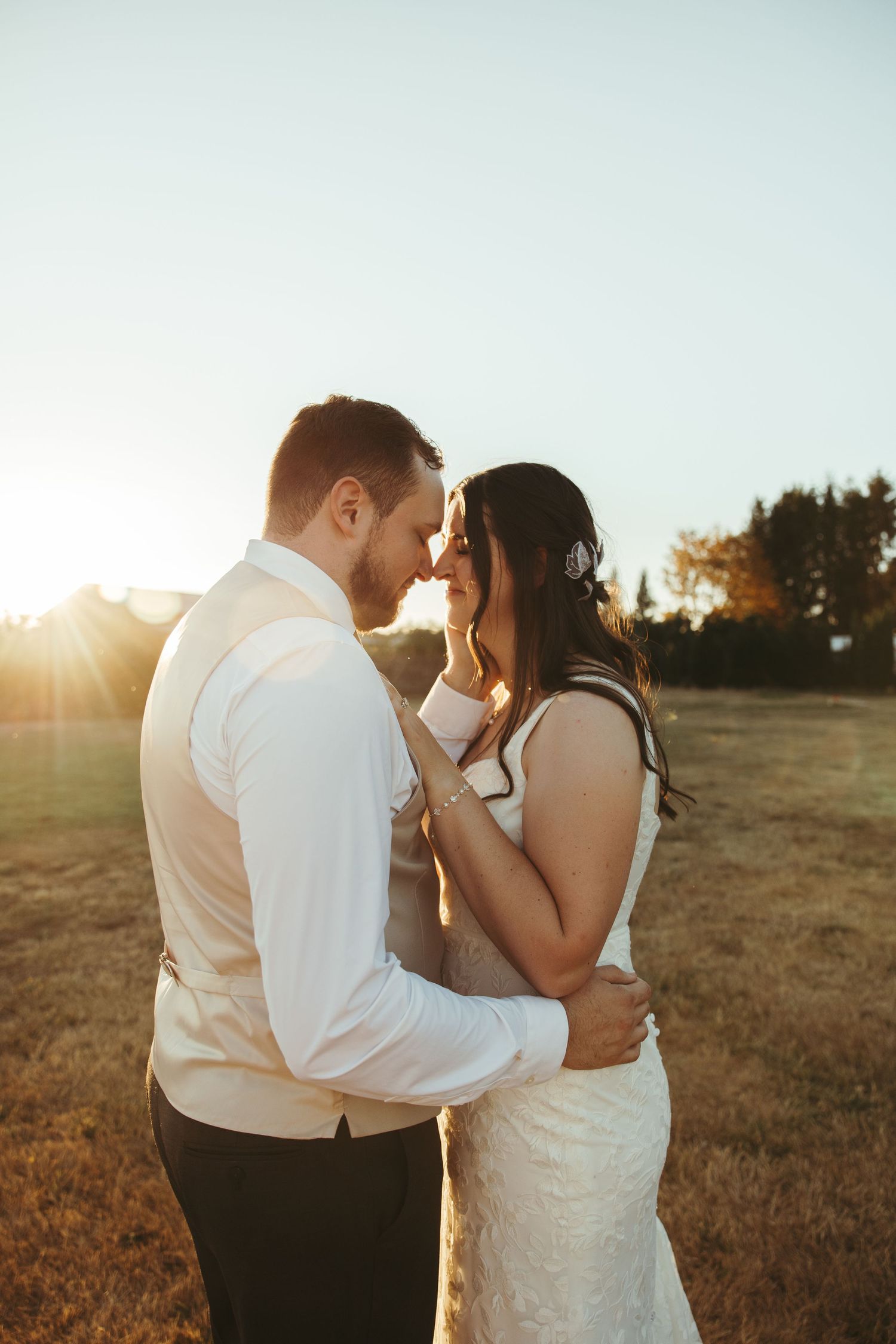 A bride and groom share a romantic forehead touch at golden hour in an open field, bathed in warm sunset light.