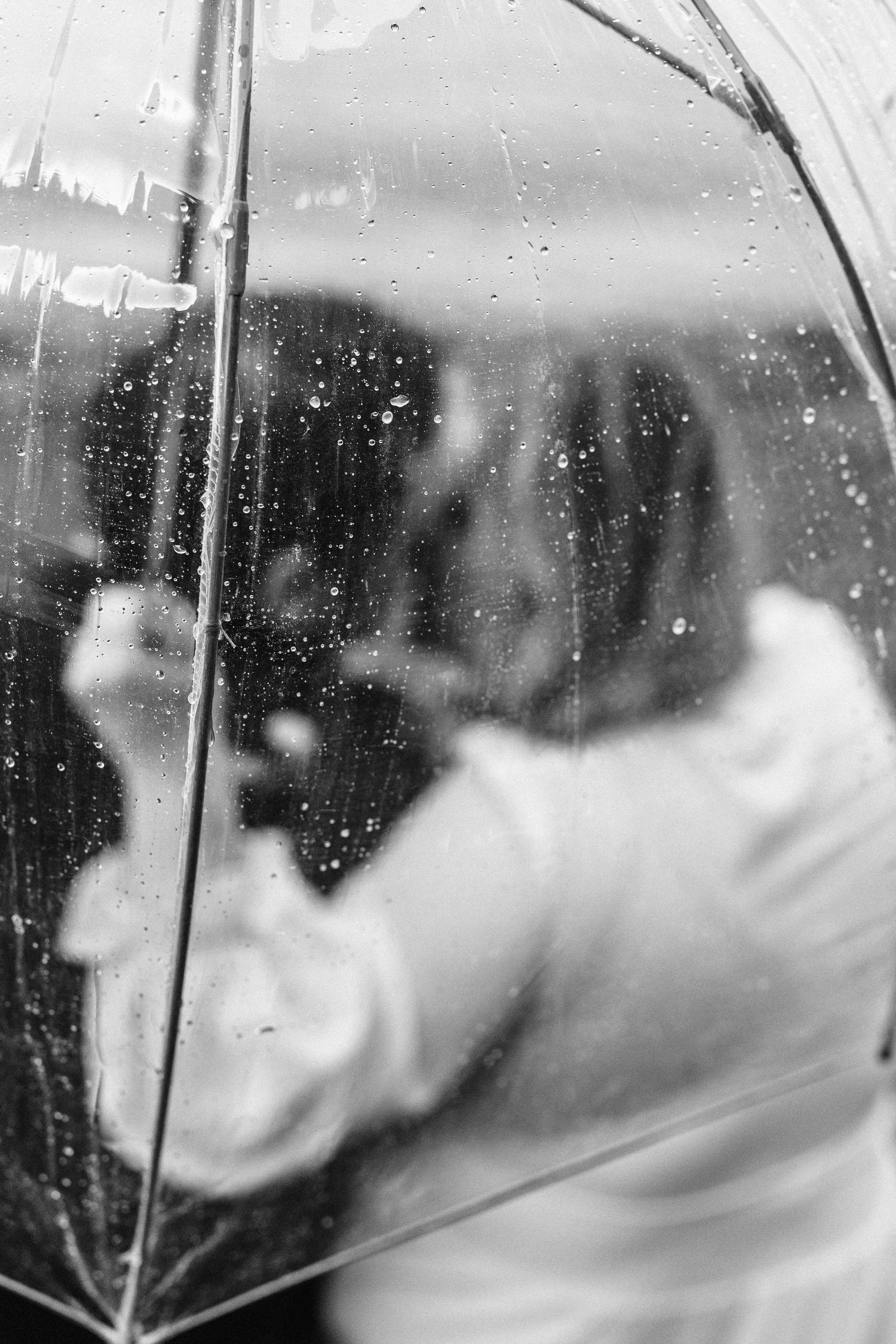 Black and white photograph of a transparent umbrella with raindrops against a blurry stormy background.