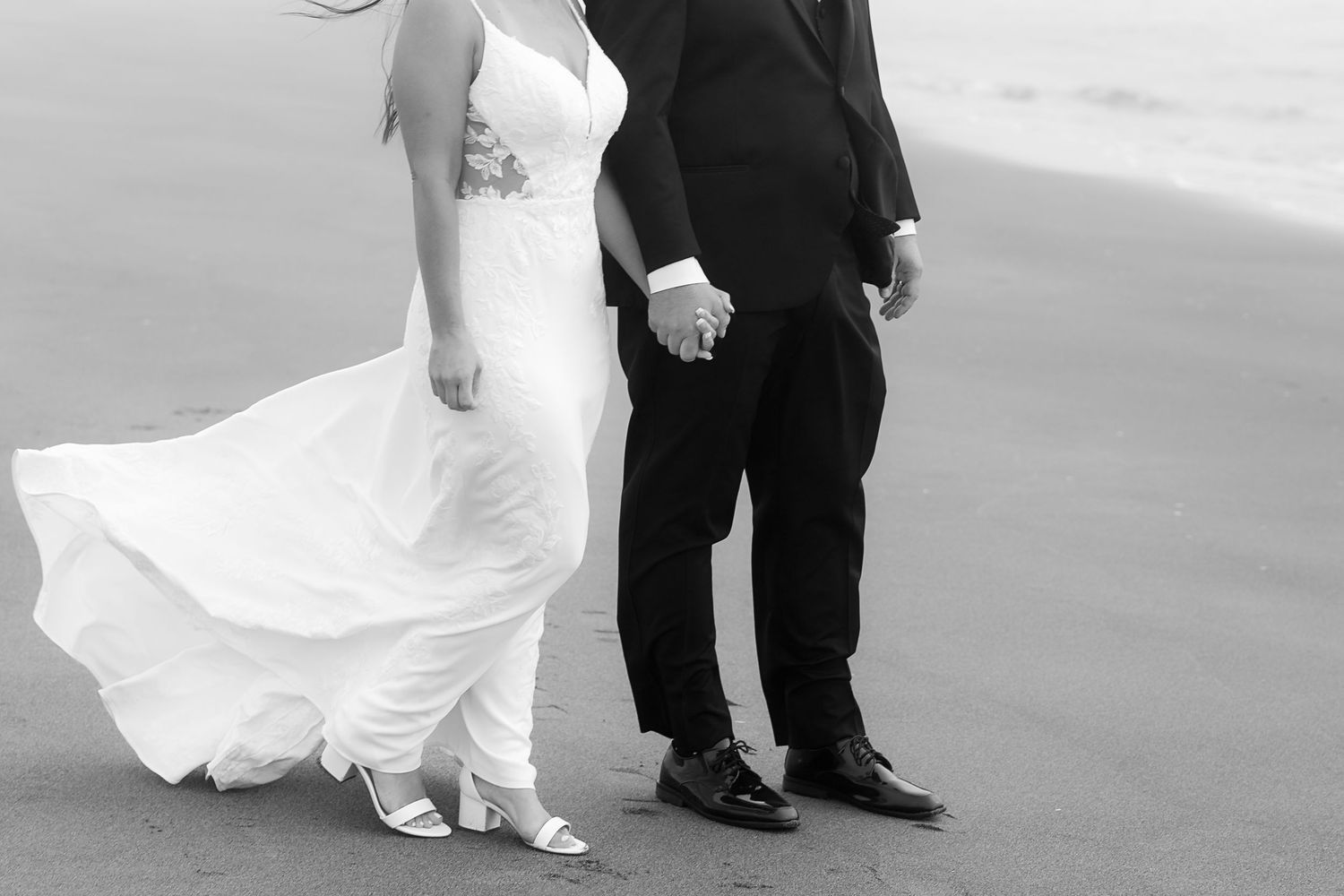 Black and white photo showing a bride in flowing white dress and groom in dark suit holding hands on beach.