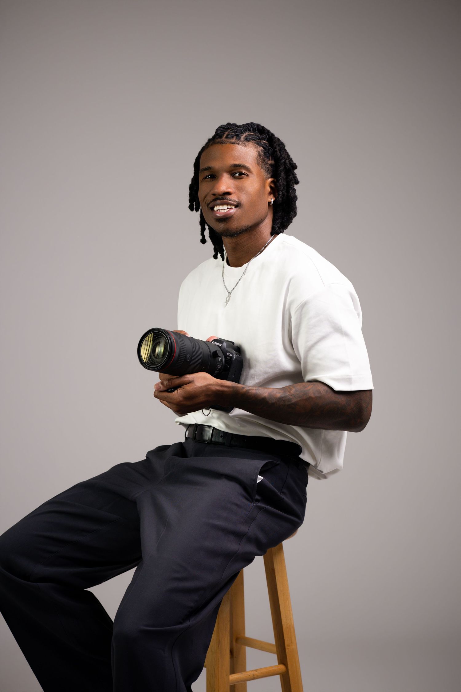 Person wearing white t-shirt and dark pants sits on wooden stool in studio setting with neutral background.