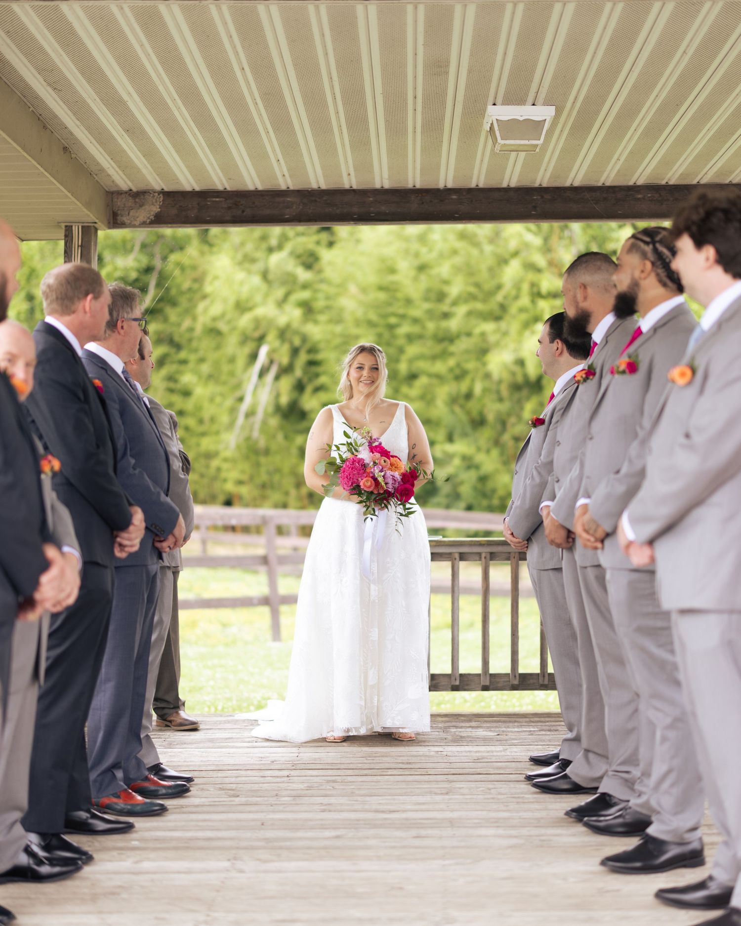 Bride in white gown holding colorful bouquet walks down aisle flanked by groomsmen in gray suits at outdoor wedding.