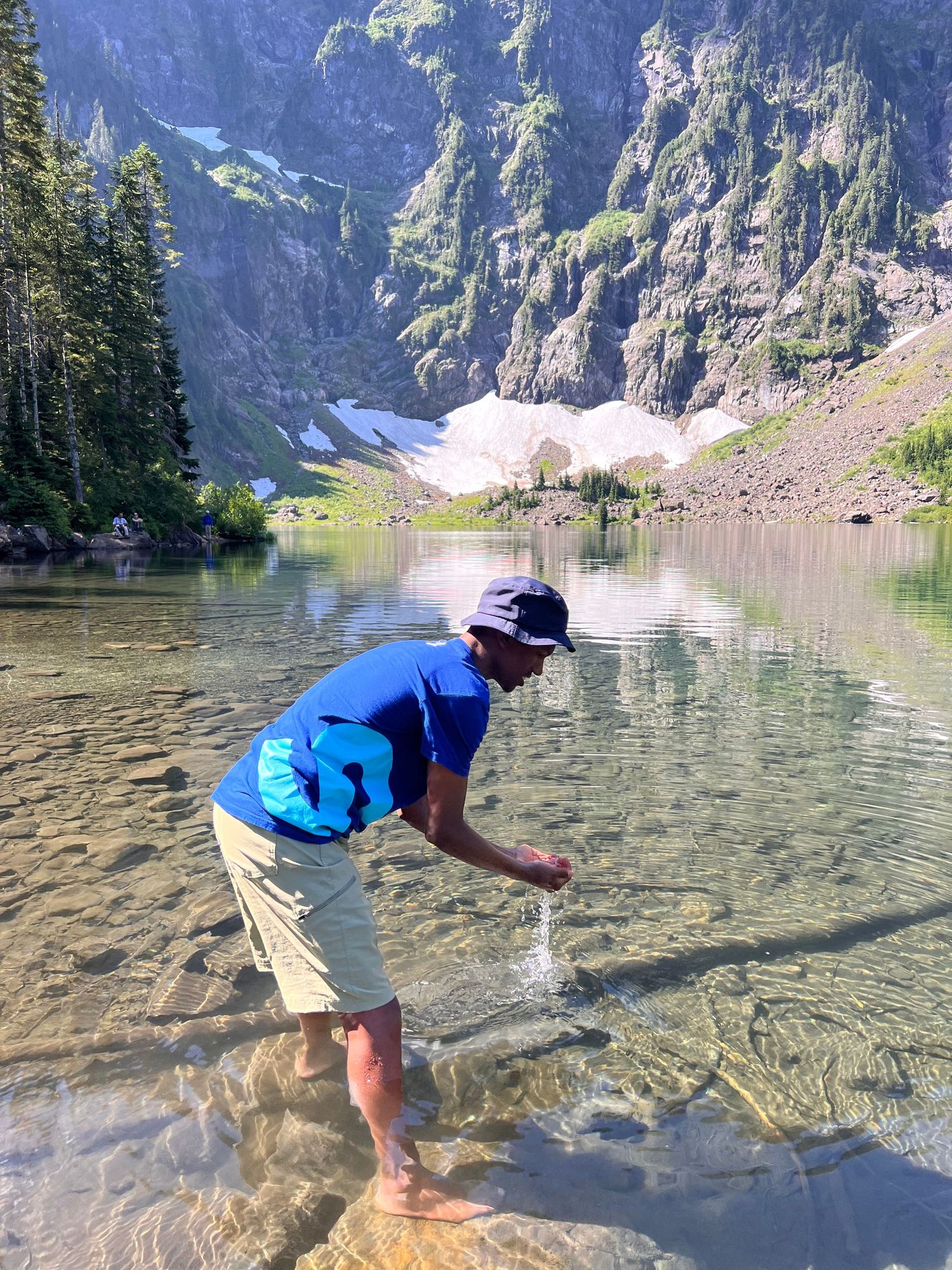 Person in blue shirt and khaki shorts bends down to touch crystal clear mountain lake water with mountain backdrop.