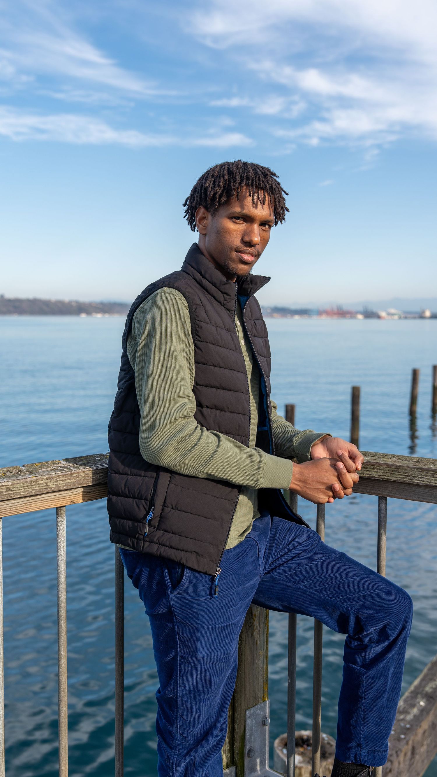 A person wearing a puffy vest and olive sweater leans on a railing overlooking a scenic waterfront view on a sunny day.