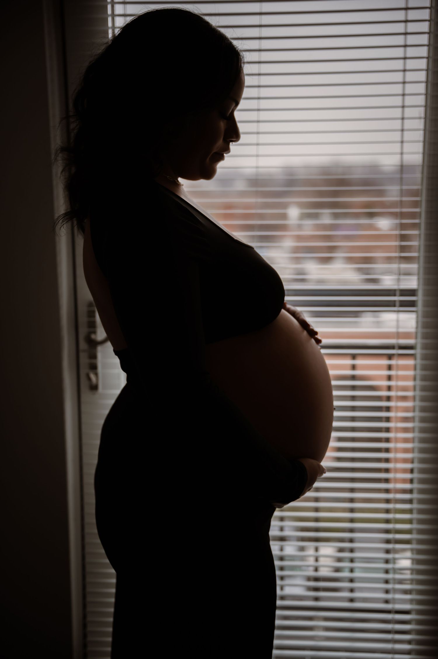 Silhouetted figure stands by window with venetian blinds casting dramatic shadows in moody maternity photo in Stafford VA.
