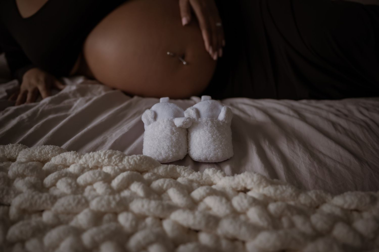 Pregnant woman sitting on bed with tiny white baby booties in foreground on textured blanket.