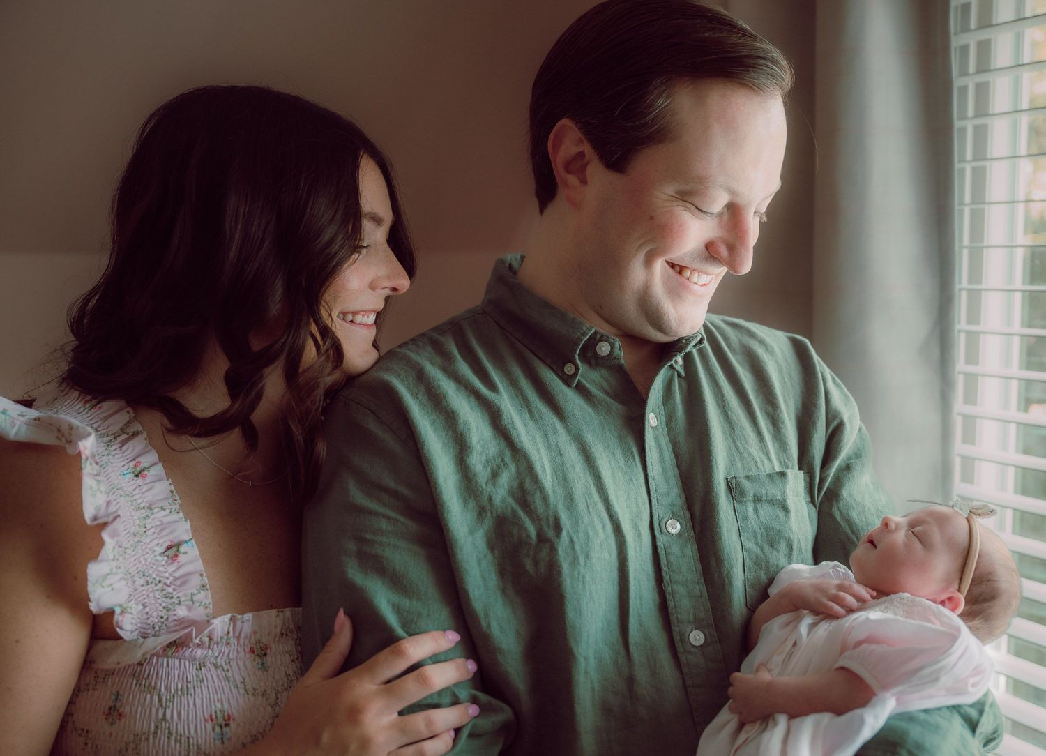 Happy couple smiling at their newborn baby by a window in a warm, intimate family portrait.