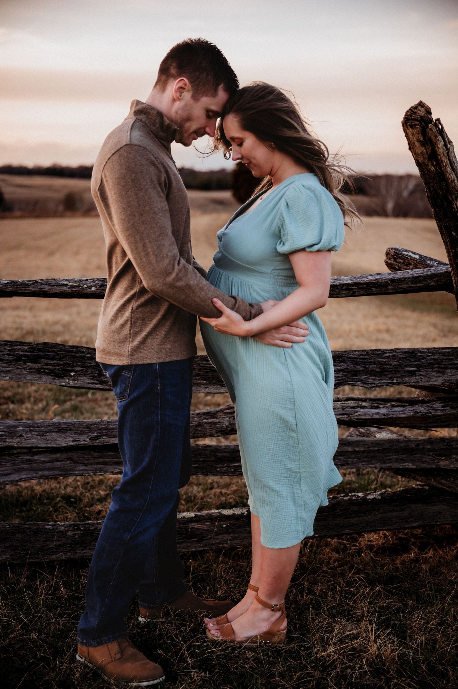 A couple shares a tender maternity moment by a rustic fence at sunset, man in brown jacket cradling pregnant womans belly.