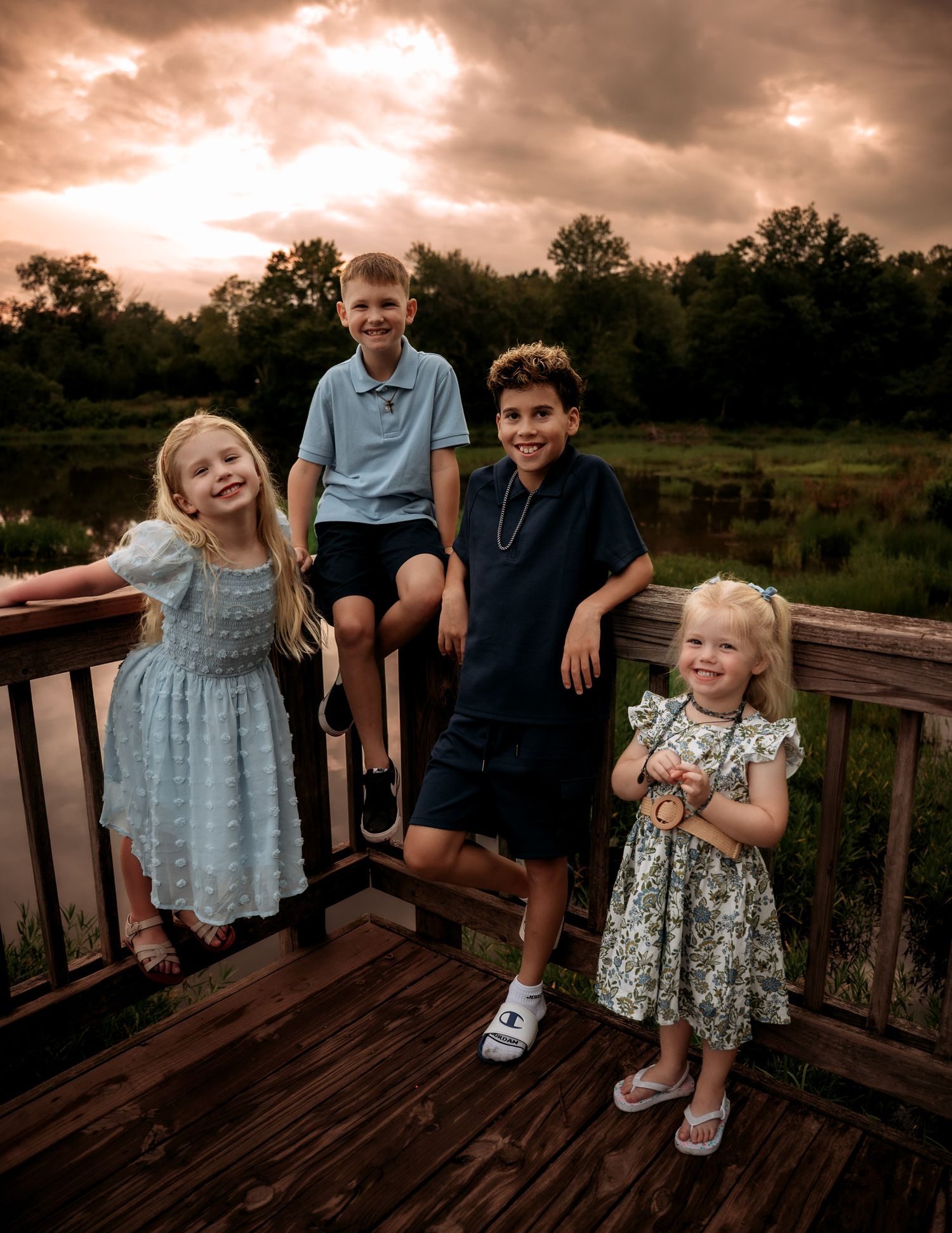 Four children posing on a wooden deck near a pond at sunset, wearing dressy outfits and smiling.