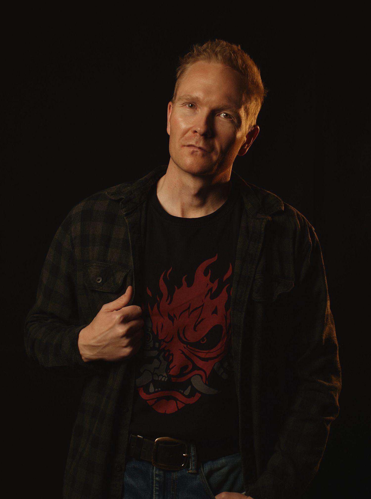 A person wearing a plaid flannel shirt and black t-shirt with red design poses against a dark background in a photo series.