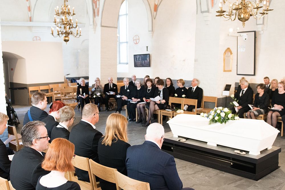 Laaja kuva hautajaisista, jossa näkyy kirkossa istuvat läheiset sekä valkoinen arkku, jonka päällä on kukkia.