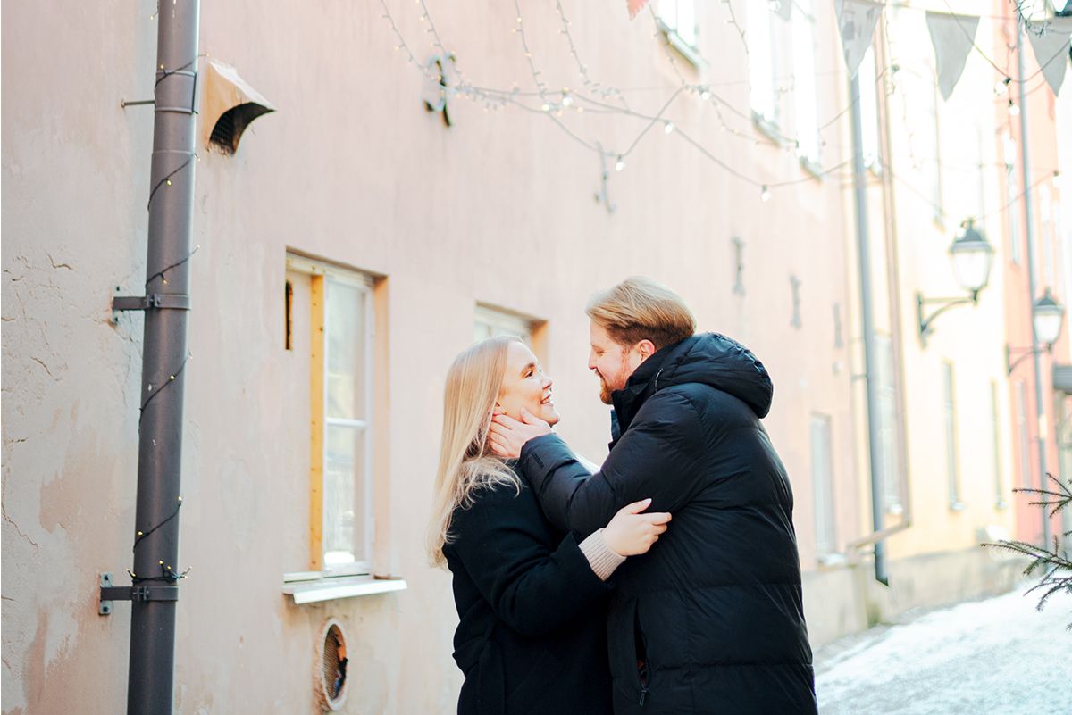 Vaaleanpunaista seinää vasten otettu pariskuntakuva, jossa mies pitää naista poskesta hellästi kiinni ja pari katsoo toisiaan lempeästi.