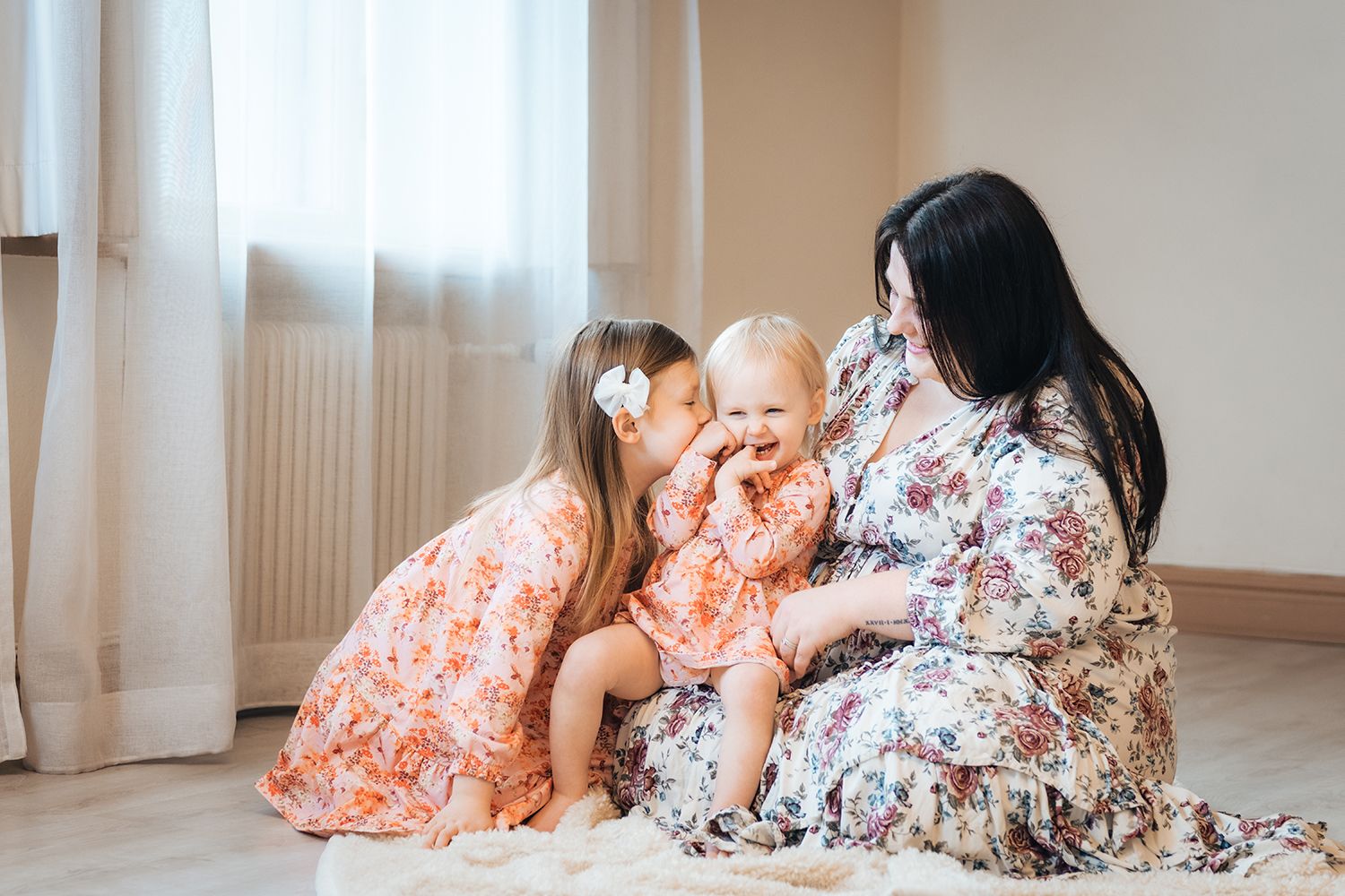 Äiti ja kaksi lasta istumassa taljan päällä lattialla ja isosisko antaa pikkusiskolleen pusua poskelle iloisesti.