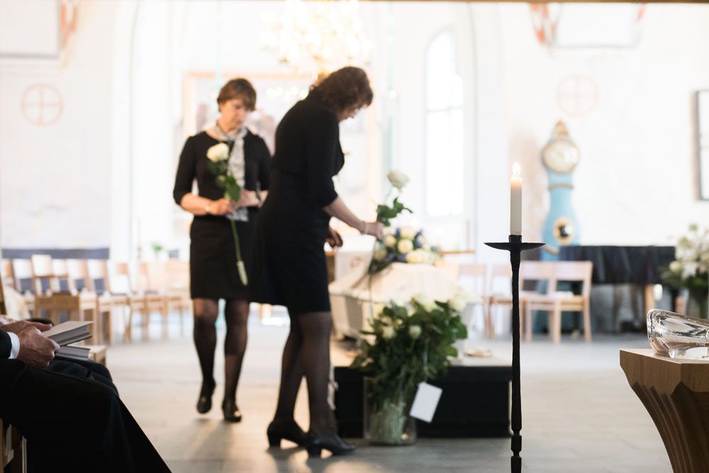 Dokumentaarinen kuva hautajaisista, jossa tarkennus on kynttilässä ja taustalla läheinen vie valkoista ruusua arkun päälle.
