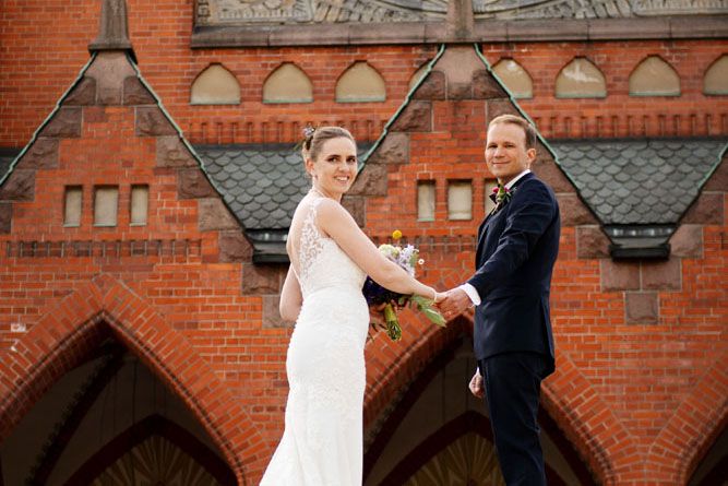 Hääpari pitää toisiaan kädestä kiinni ja katsoo olan yli kameraan. Heidän takanaan näkyy Mikaelin kirkko.