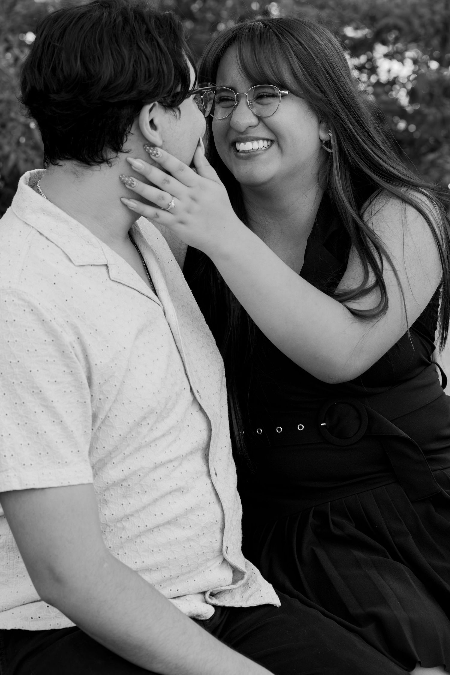 Black and white photo of a smiling woman with glasses touching a man's face as they sit together outdoors.