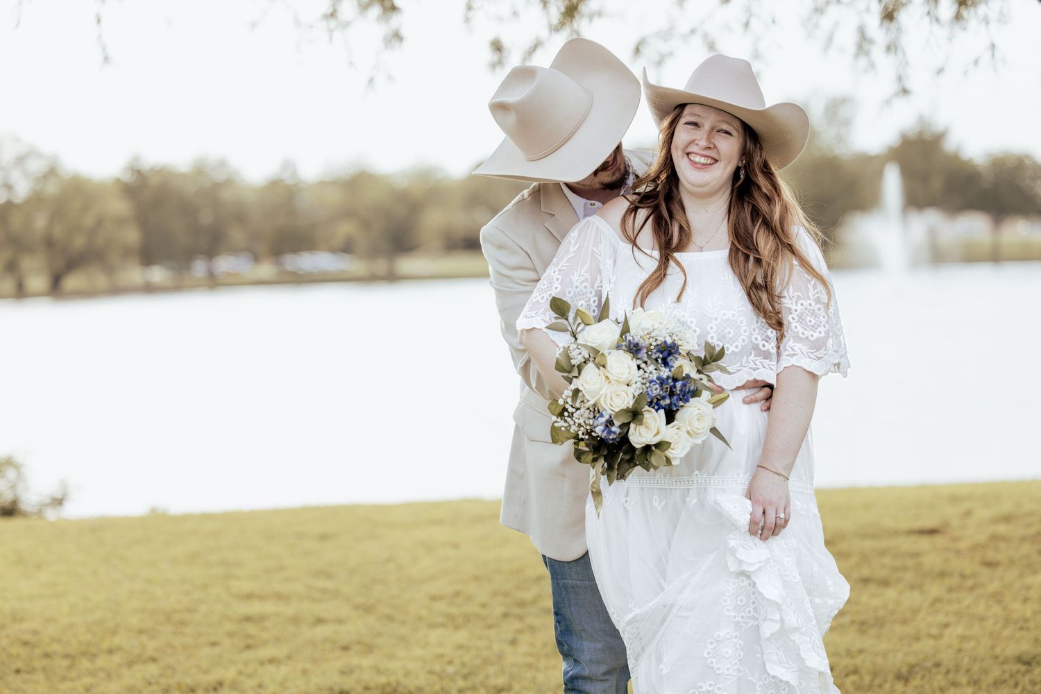 Bride in white dress and cowboy hat holds bouquet while groom kisses her cheek near a lake.