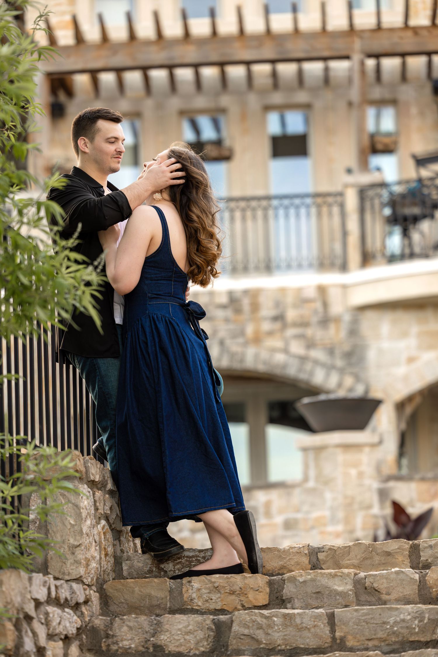 Romantic couple embracing on stone steps, woman in blue dress, man in black jacket, elegant building backdrop.