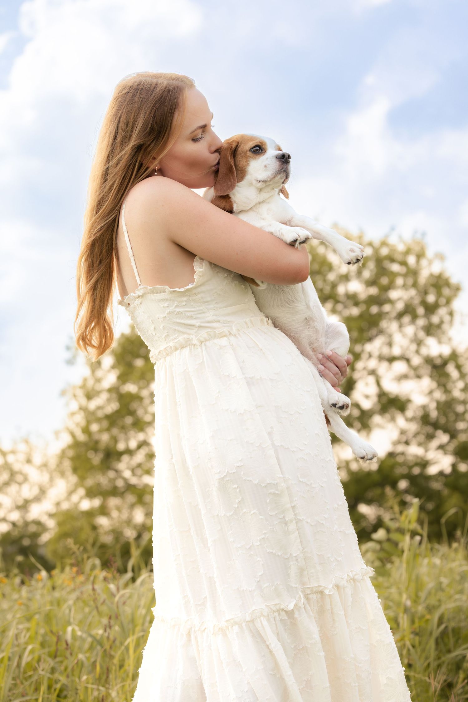 Young woman in white dress kissing her Jack Russell terrier dog outdoors in a green field.