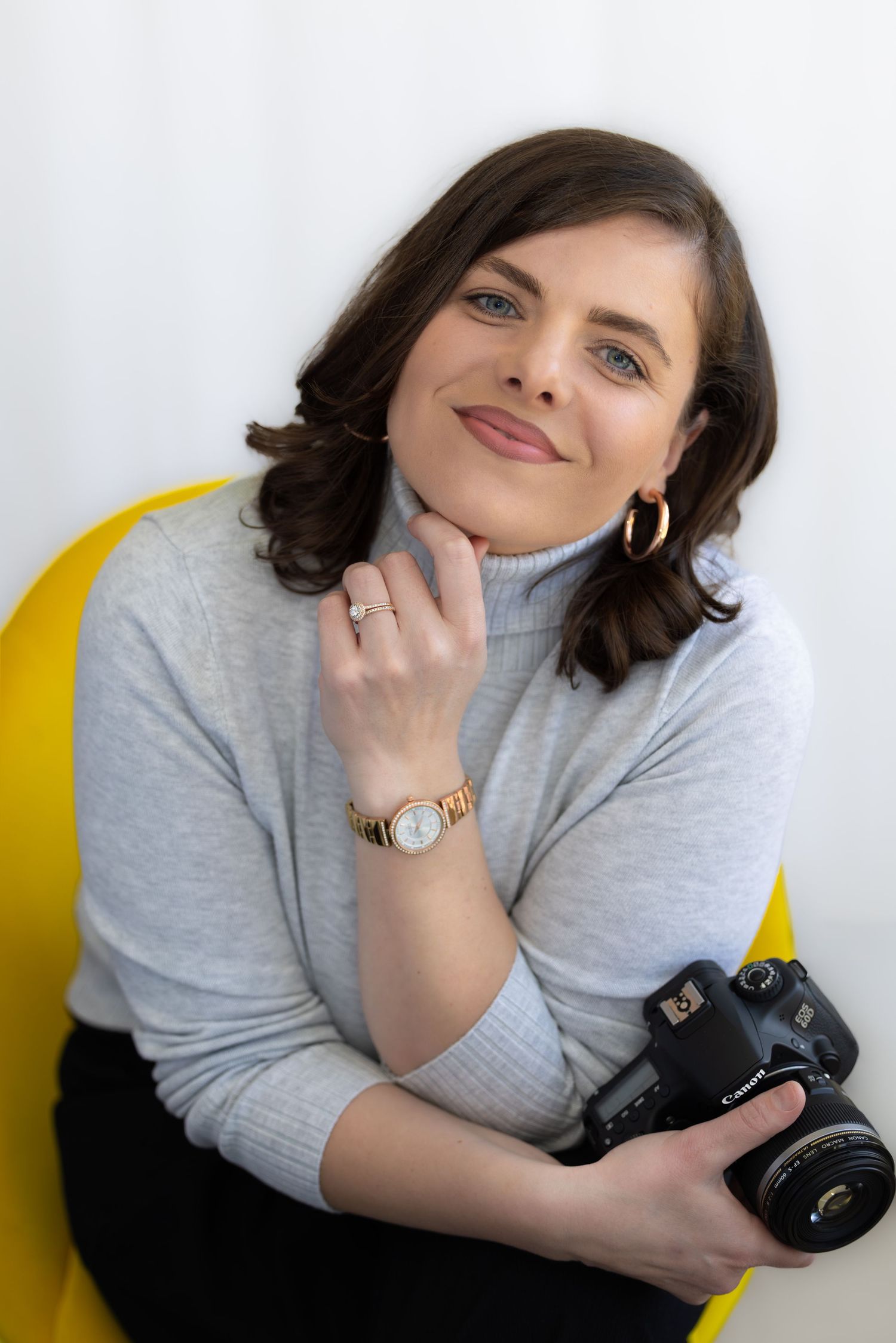 A photographer in a gray turtleneck sweater poses with a DSLR camera while sitting in a yellow chair.