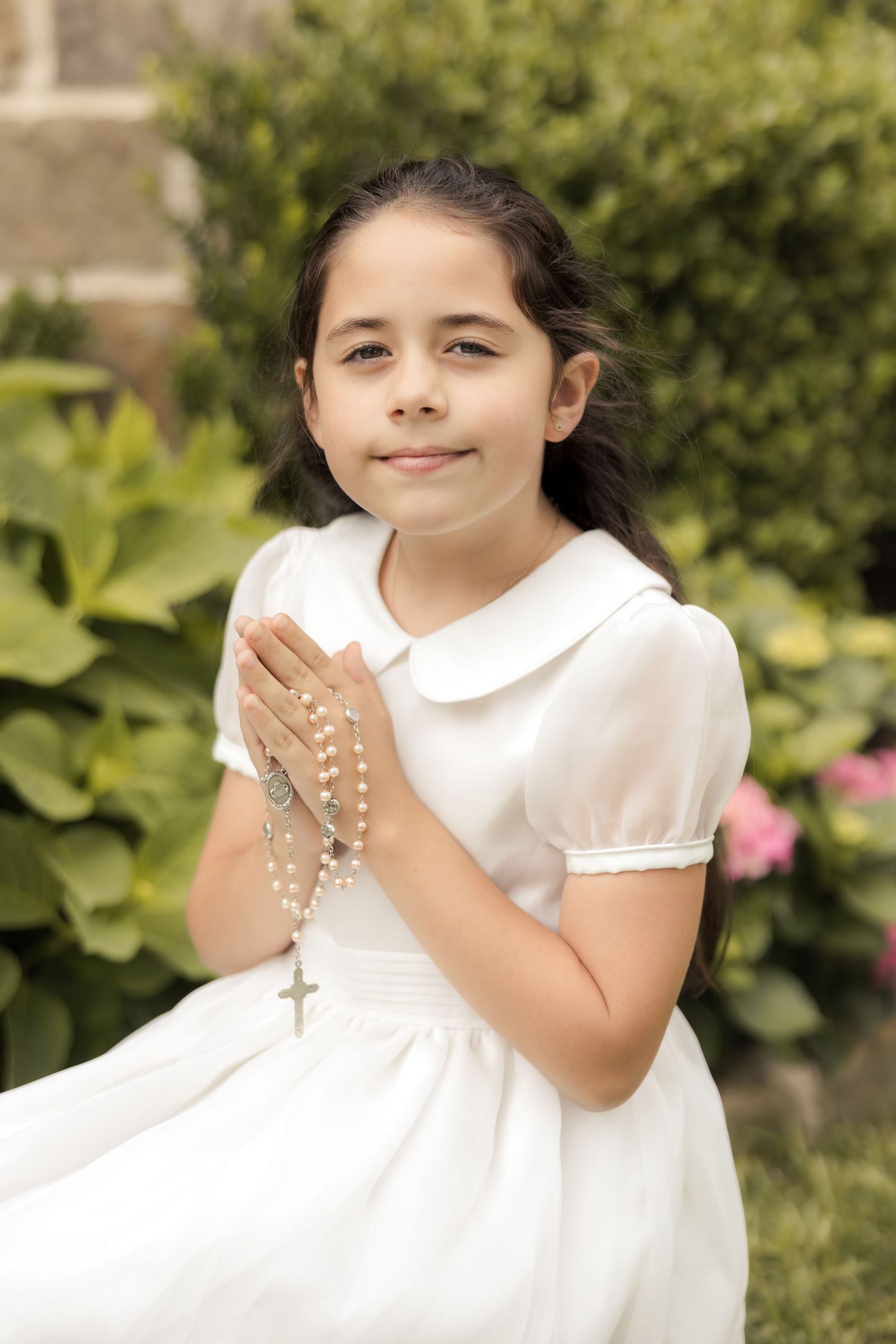 Young girl in white dress holding rosary beads in prayer pose, celebrating First Communion outdoors.